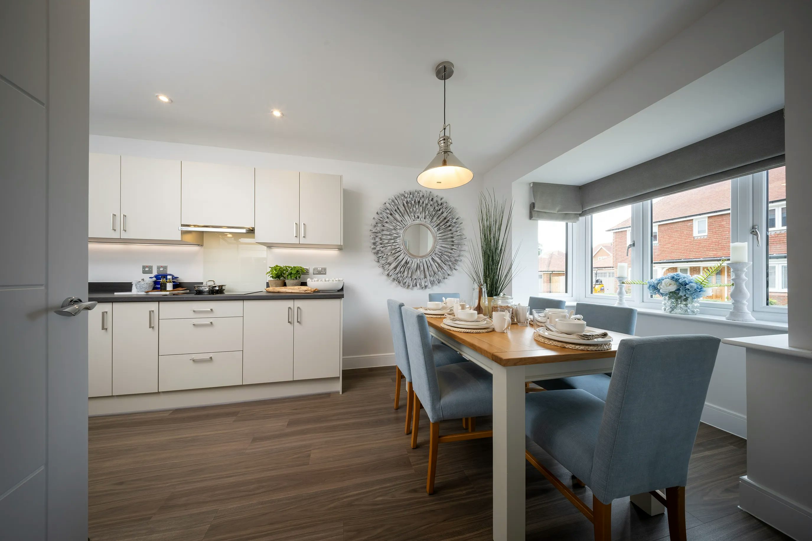 A modern kitchen and dining area with white cabinets, a stove with a pot, and a countertop featuring various items like a plant and a cutting board. The dining space includes a wooden table set with plates, bowls, cups, and cutlery, surrounded by four upholstered chairs. A decorative mirror hangs on the wall, while a pendant light illuminates the table. Large windows let in natural light and reveal a view of neighboring houses. The room has wooden flooring and a clean, contemporary design.