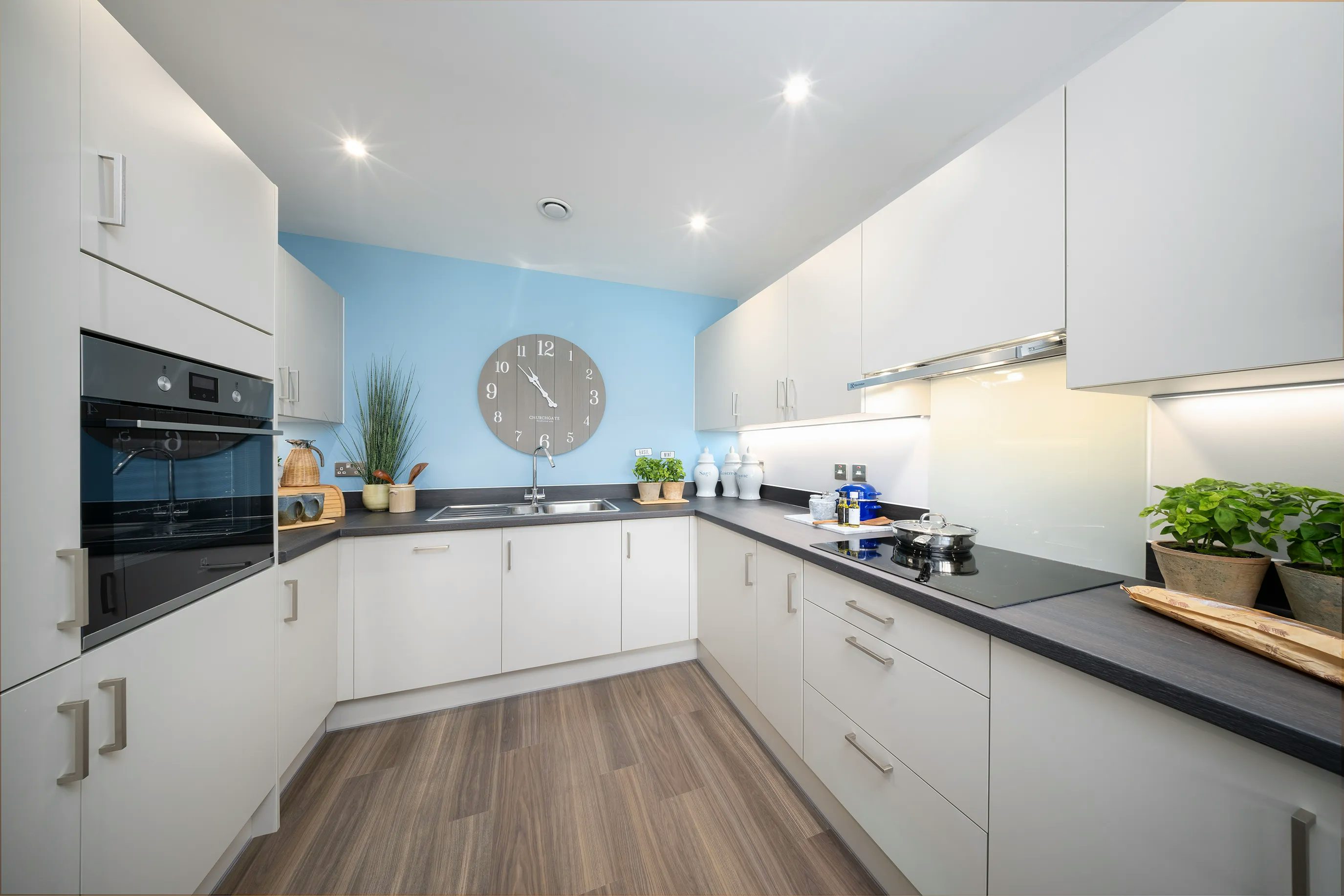 A modern kitchen with white cabinetry and a light blue accent wall. The kitchen is well-organized, featuring a built-in oven, stovetop with pots and pans, a sink, and various kitchen accessories, including cutting boards, plants, and decorative items. A large round clock hangs above the sink. The space has ample counter space and lighting, creating a bright and functional atmosphere.