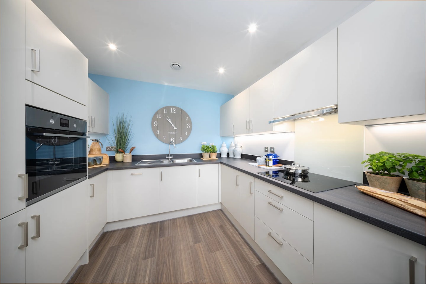 A modern kitchen with white cabinetry and a light blue accent wall. The kitchen is well-organized, featuring a built-in oven, stovetop with pots and pans, a sink, and various kitchen accessories, including cutting boards, plants, and decorative items. A large round clock hangs above the sink. The space has ample counter space and lighting, creating a bright and functional atmosphere.