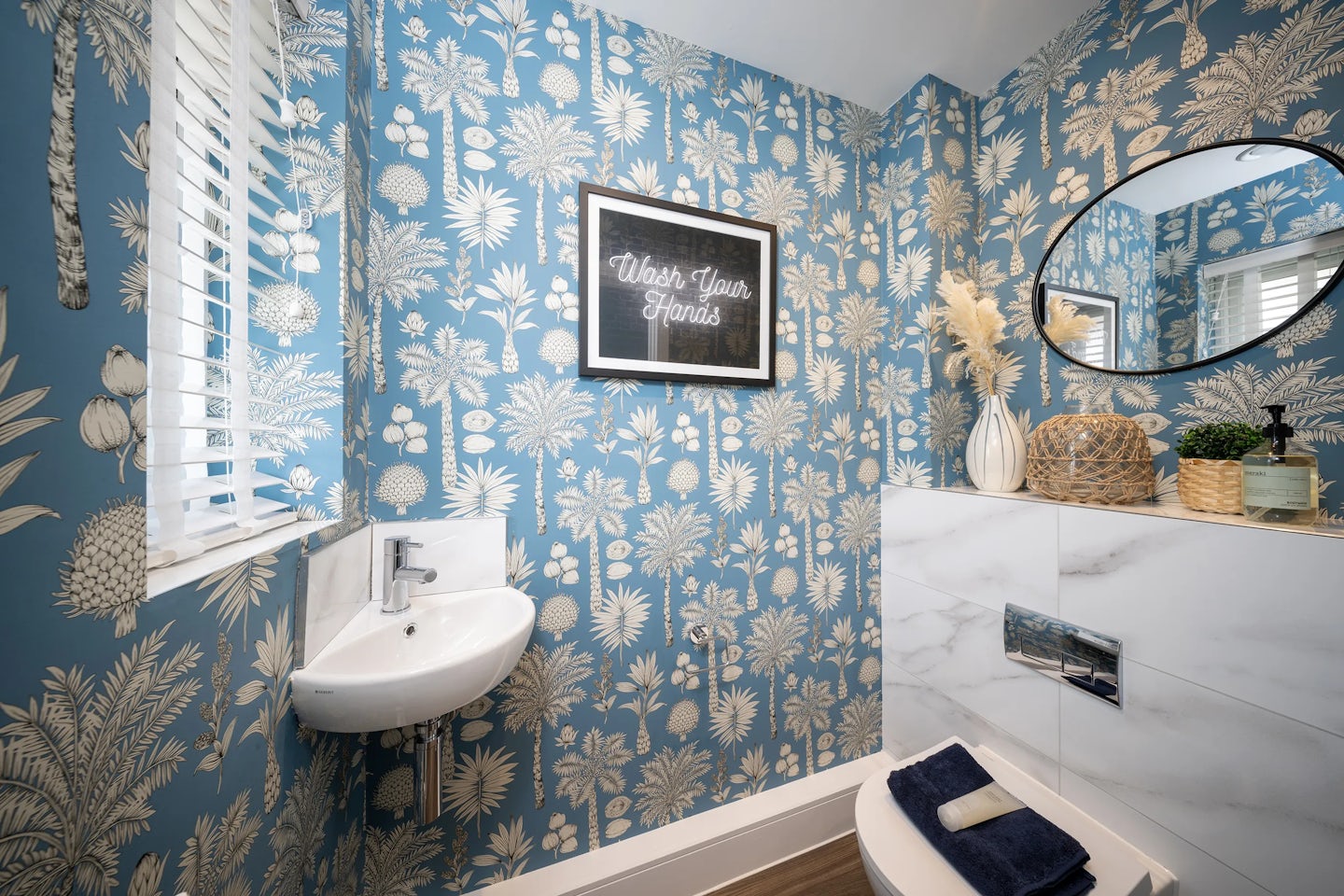 A small, stylish bathroom featuring blue wallpaper with white botanical patterns. A compact white sink is mounted on the wall, with a round mirror above a shelf holding decorative items like a wicker basket, a small plant, and a vase with dried flowers. The toilet has a neatly folded towel on top, and a framed sign reading "Wash Your Hands" is displayed on the wall. A window with white blinds allows natural light to brighten the space.