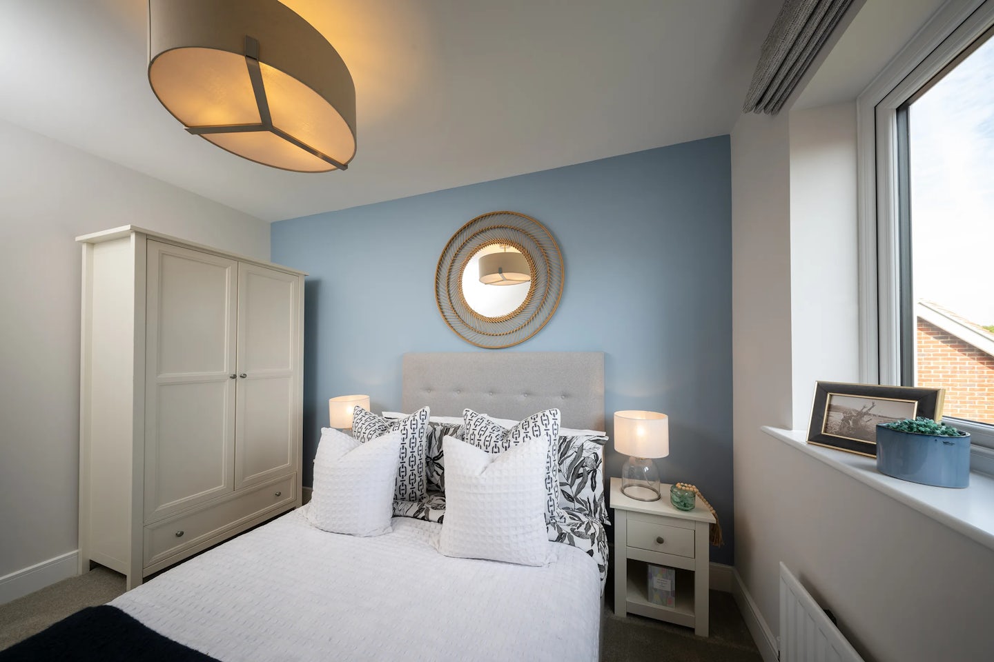 A modern bedroom with a blue accent wall behind a neatly made bed with white bedding and multiple pillows. Above the bed hangs a large circular mirror with a decorative frame. A nightstand beside the bed holds a lamp and small decorative items. The room includes a wardrobe and a ceiling light fixture. A large window allows natural light to illuminate the space, creating a cozy and inviting atmosphere. The furniture and layout are well-organized, giving the room a tidy and stylish appearance.