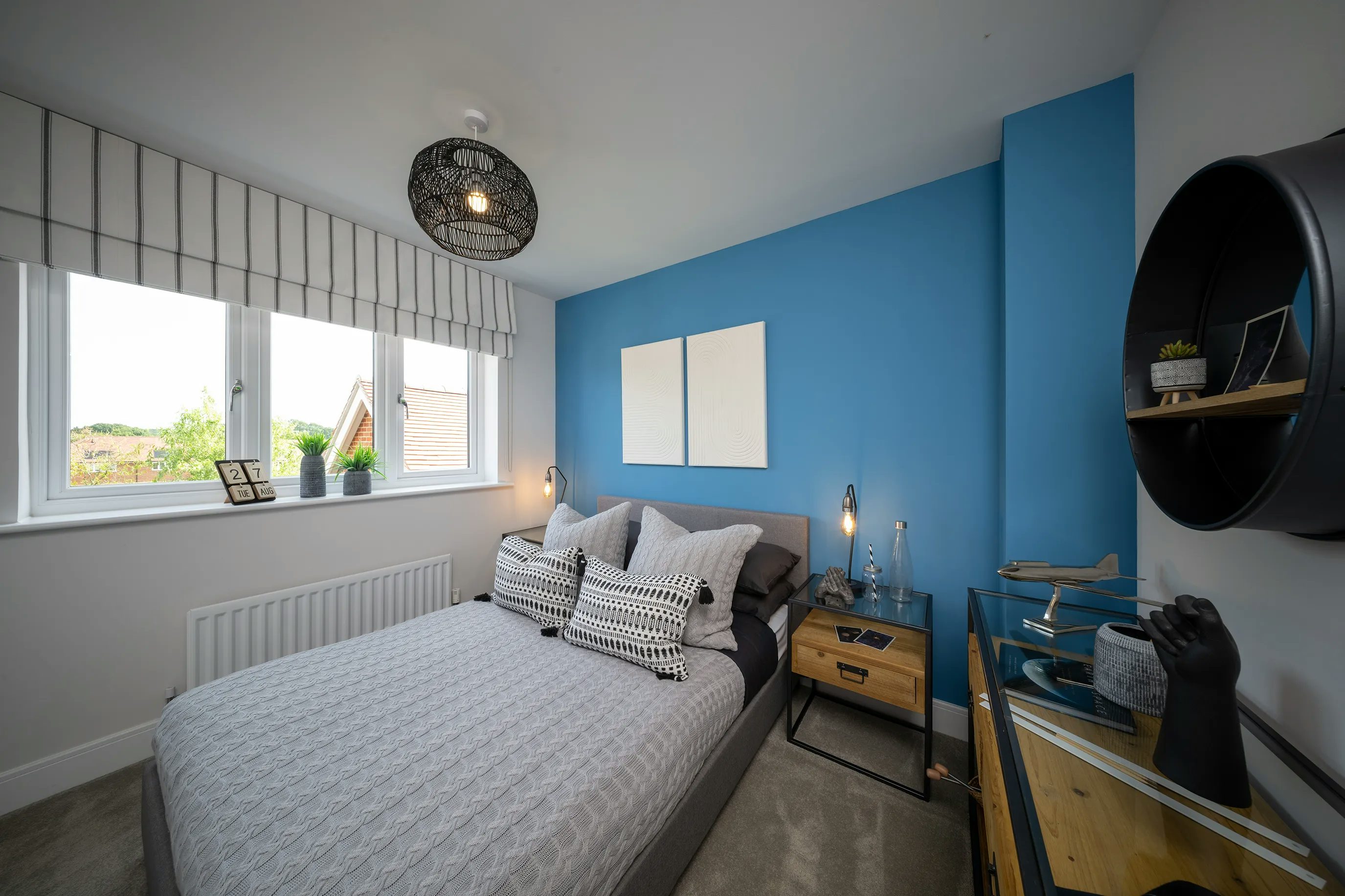 A modern bedroom with a grey quilted bedspread on a central bed, accompanied by decorative pillows in black and white patterns. The wall behind the bed is painted deep blue, featuring two square artworks above the headboard. Two bedside tables flank the bed, each holding lamps and decorative objects. A large window with a striped valance allows natural light to filter in. A black circular shelf on the right side holds various items, including a small plant. The room’s decor blends contemporary styling with cozy elements.