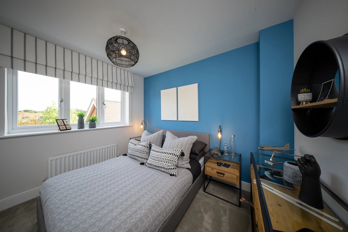 A modern bedroom with a grey quilted bedspread on a central bed, accompanied by decorative pillows in black and white patterns. The wall behind the bed is painted deep blue, featuring two square artworks above the headboard. Two bedside tables flank the bed, each holding lamps and decorative objects. A large window with a striped valance allows natural light to filter in. A black circular shelf on the right side holds various items, including a small plant. The room’s decor blends contemporary styling with cozy elements.
