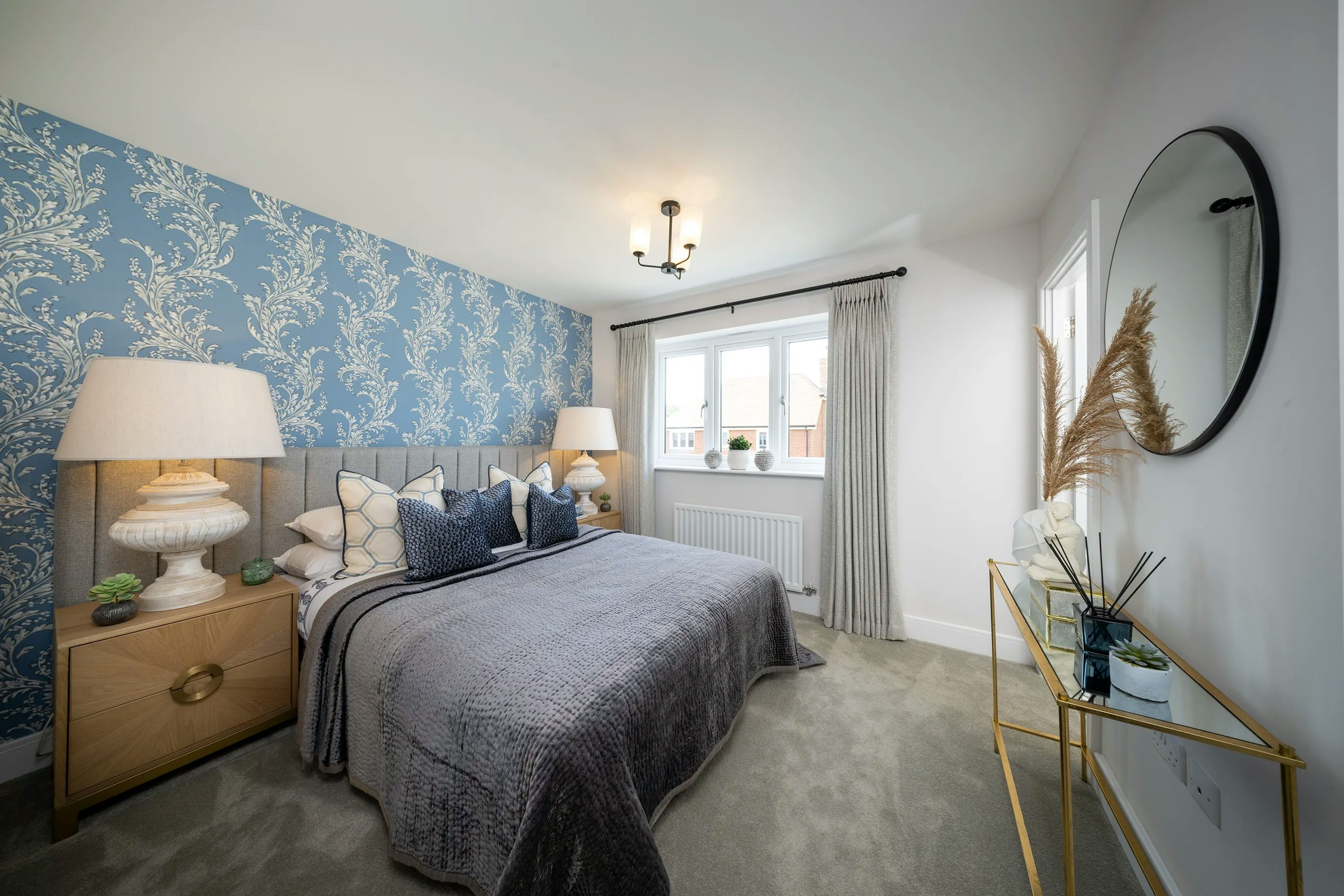 A bedroom with a contemporary design, featuring a double bed adorned with a grey quilt and an assortment of decorative pillows, including two large dark blue pillows and several smaller patterned ones. The bed is flanked by wooden nightstands, each holding a lamp and decorative accents. The wall behind the bed is covered in blue wallpaper with a white floral pattern, adding elegance. A window with grey curtains allows natural light to enter, with a radiator positioned below. To the right, a glass console table with a gold frame holds vases, dried plants, and other decorative items. Above it, a round mirror reflects the room’s soft lighting. The ceiling light fixture contributes to the warm ambiance. Would you like any refinements to better align with your accessibility or branding needs?
