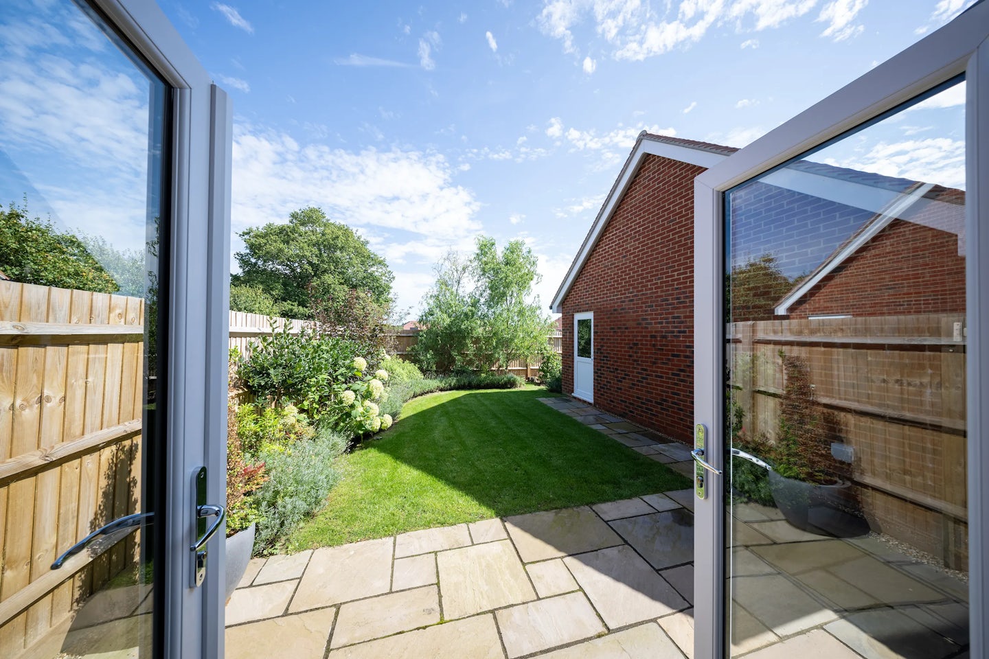 A view of a backyard garden seen through open double glass doors from inside a house. The garden features a neatly trimmed lawn bordered by a variety of plants and shrubs. A wooden fence runs along the left side, while a brick outbuilding, possibly a garage or shed, stands on the right. The sky is bright with scattered clouds, suggesting a pleasant day. The scene conveys a sense of suburban tranquillity and well-maintained outdoor space.