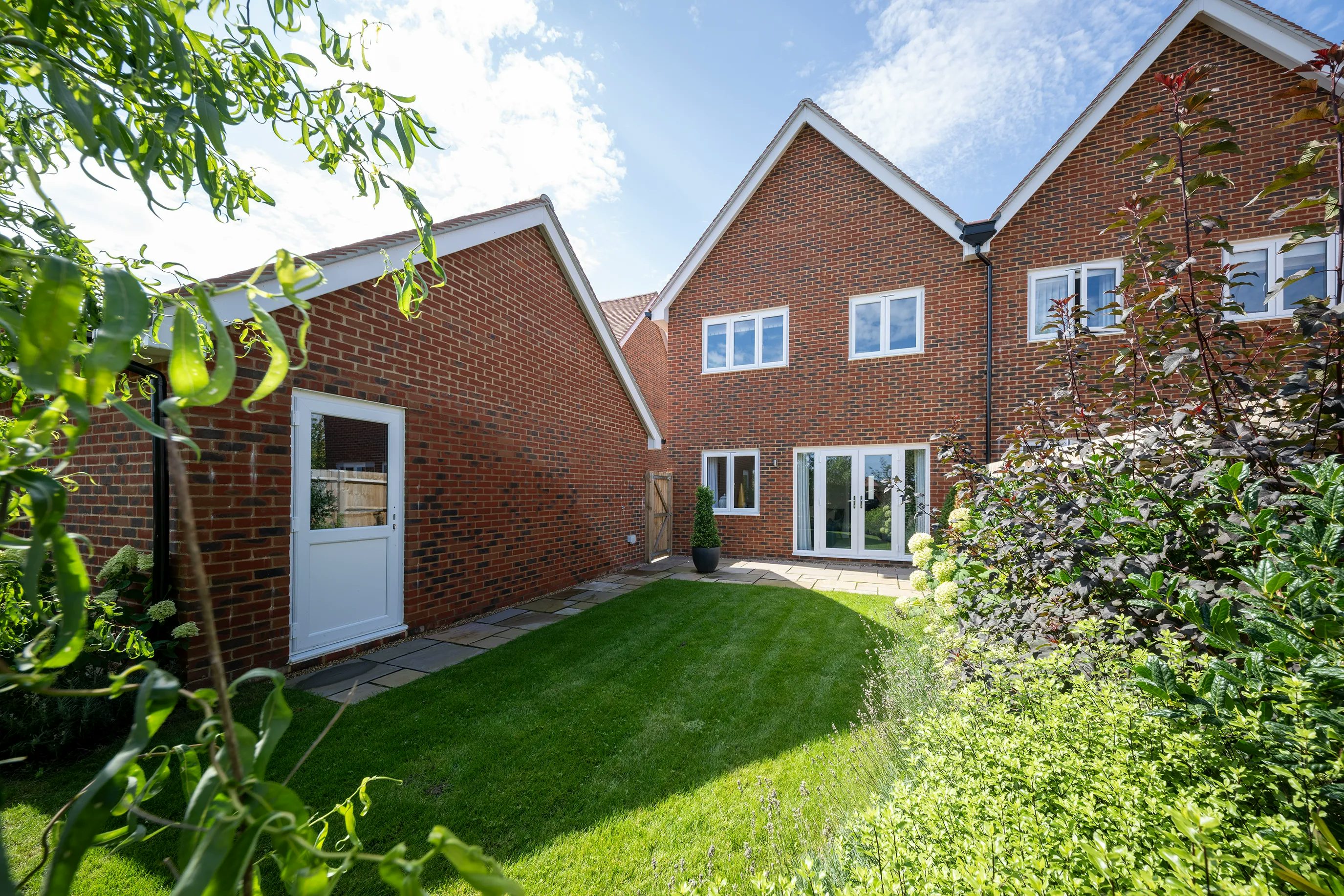 A suburban backyard with a neatly trimmed lawn and landscaped garden. The house, made of red bricks with a pitched roof, features two visible sections—one with a white door and another with large glass doors and windows. Various plants and shrubs surround the outdoor space, complementing the home’s tidy aesthetic. The sky is partly cloudy, casting soft natural light over the scene.