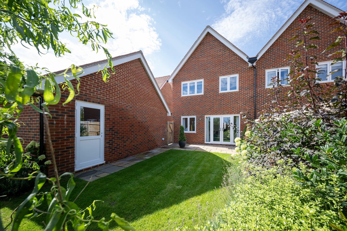 A suburban backyard with a neatly trimmed lawn and landscaped garden. The house, made of red bricks with a pitched roof, features two visible sections—one with a white door and another with large glass doors and windows. Various plants and shrubs surround the outdoor space, complementing the home’s tidy aesthetic. The sky is partly cloudy, casting soft natural light over the scene.