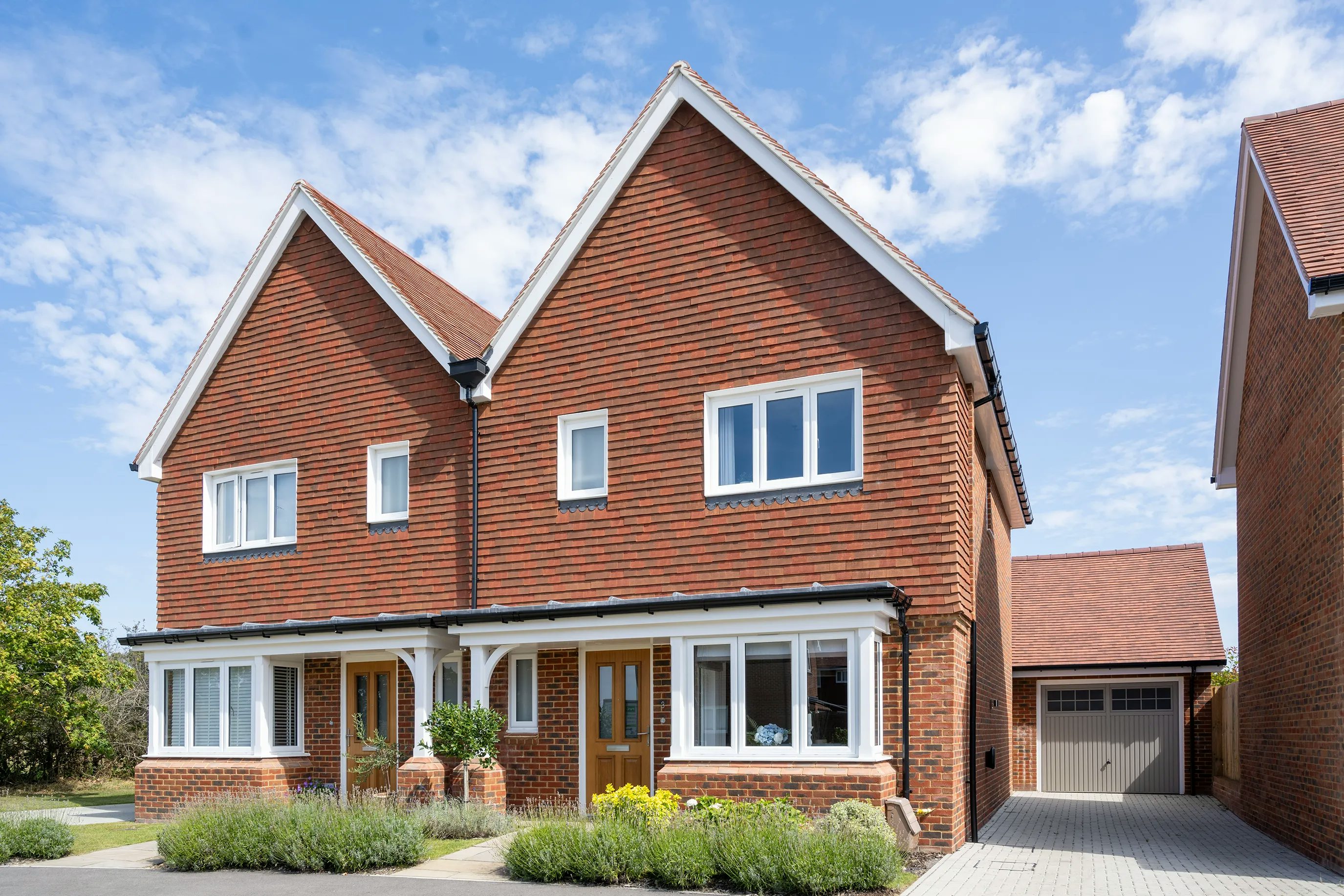 A pair of semi-detached houses with red brick exteriors, white-framed windows, and symmetrical gabled roofs. Each house features a wooden front door with a small porch area and greenery in front. A shared central wall divides them, reinforcing their mirrored design. A driveway leads to a garage on the right side. The clear sky with a few clouds suggests a bright and pleasant day.