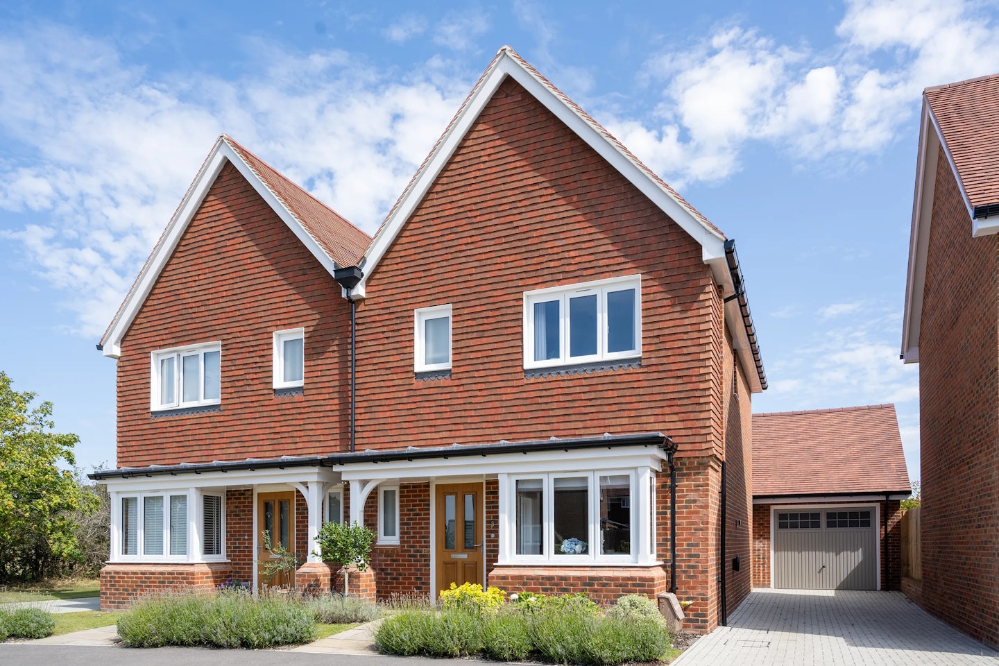 A pair of semi-detached houses with red brick exteriors, white-framed windows, and symmetrical gabled roofs. Each house features a wooden front door with a small porch area and greenery in front. A shared central wall divides them, reinforcing their mirrored design. A driveway leads to a garage on the right side. The clear sky with a few clouds suggests a bright and pleasant day.