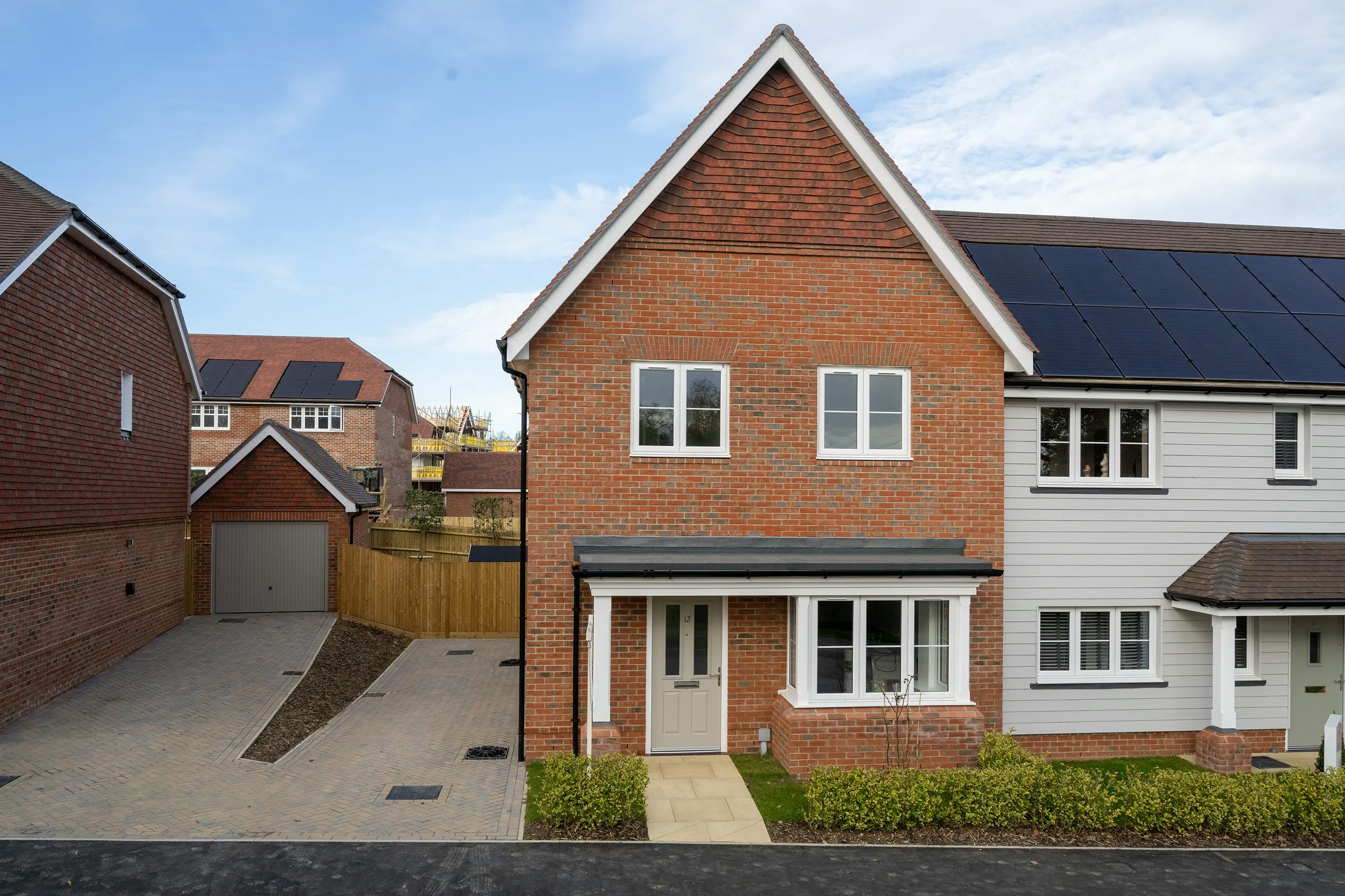 A modern suburban home with a brick facade and a grey front door. The two-story house features solar panels on its roof, indicating an environmentally friendly design. A paved pathway leads through a small front garden to the entrance. To the left, a driveway provides access to a detached garage. The surrounding neighborhood consists of similarly styled homes, creating a cohesive architectural aesthetic.