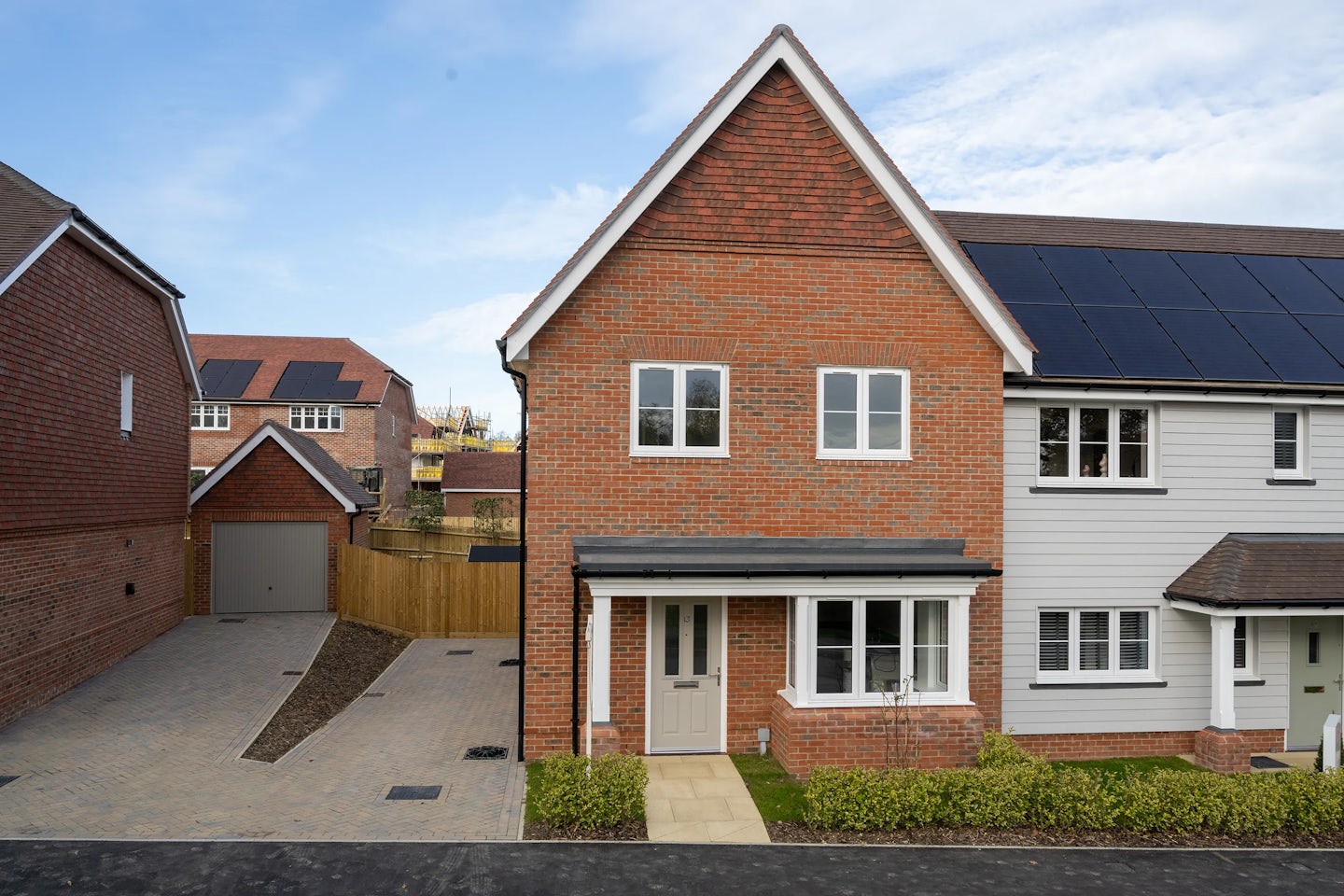 A modern suburban home with a brick facade and a grey front door. The two-story house features solar panels on its roof, indicating an environmentally friendly design. A paved pathway leads through a small front garden to the entrance. To the left, a driveway provides access to a detached garage. The surrounding neighborhood consists of similarly styled homes, creating a cohesive architectural aesthetic.