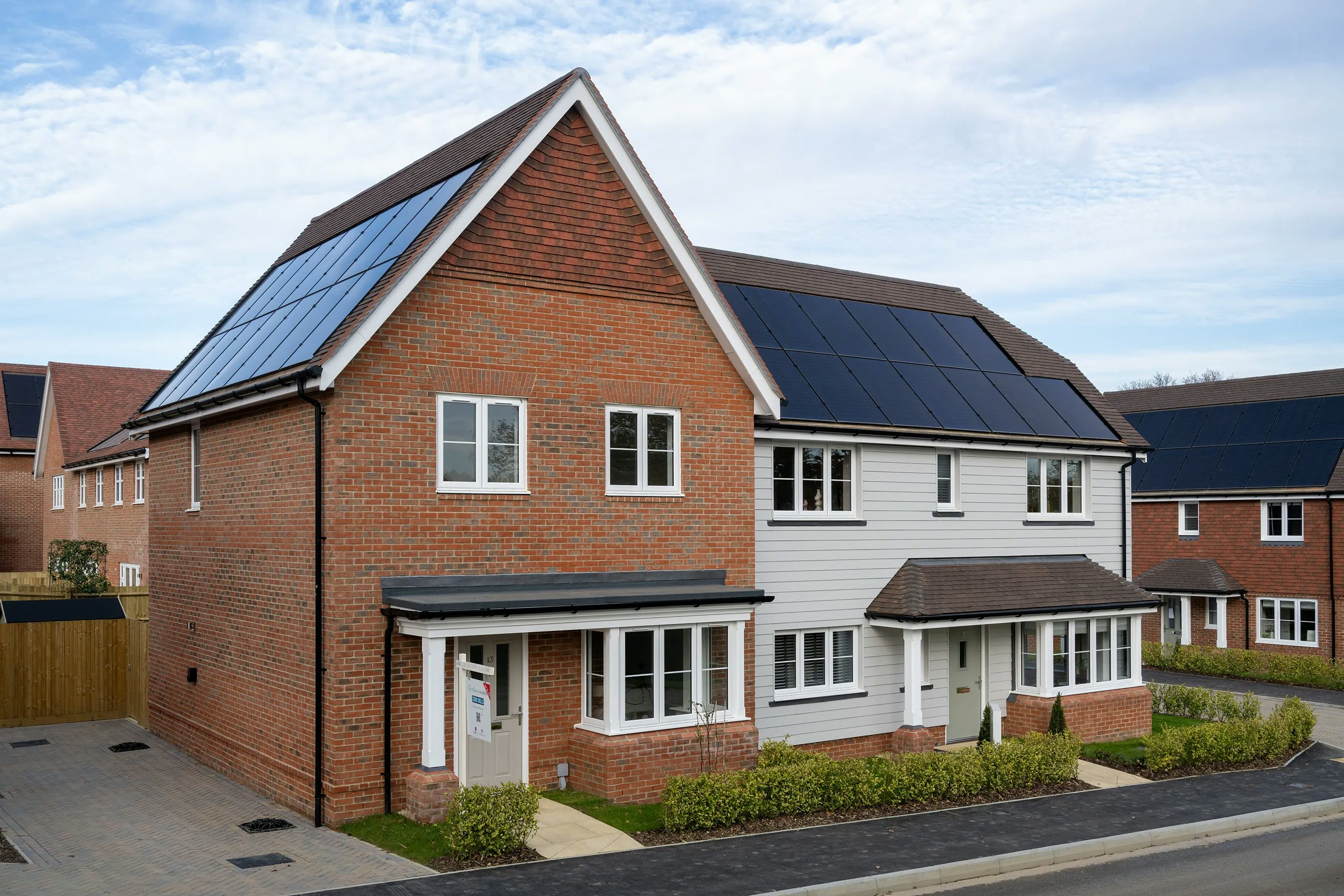 A modern suburban home with a pitched roof fitted with solar panels. The exterior combines brick and white siding, with multiple windows enhancing natural light. A neatly landscaped front garden adds to the welcoming aesthetic. The presence of solar panels suggests a focus on renewable energy and sustainability.