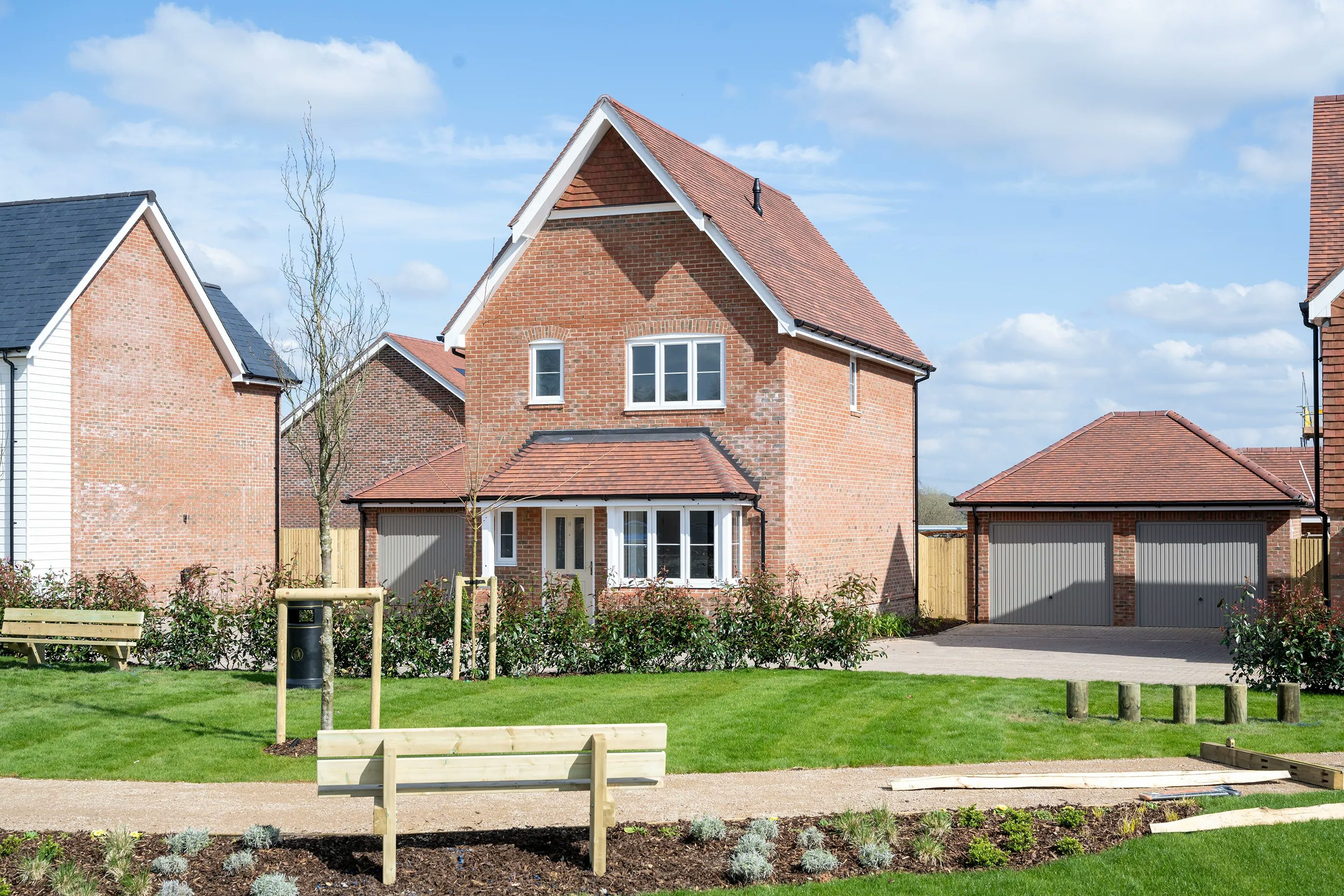 A modern two-story brick house with a gable roof, featuring a detached double garage. The home is set within a well-maintained front yard, with lush green grass, a small tree, and a pathway leading to the front door. In the foreground, benches and landscaped areas contribute to the suburban aesthetic. The sky is partly cloudy, suggesting a pleasant day.