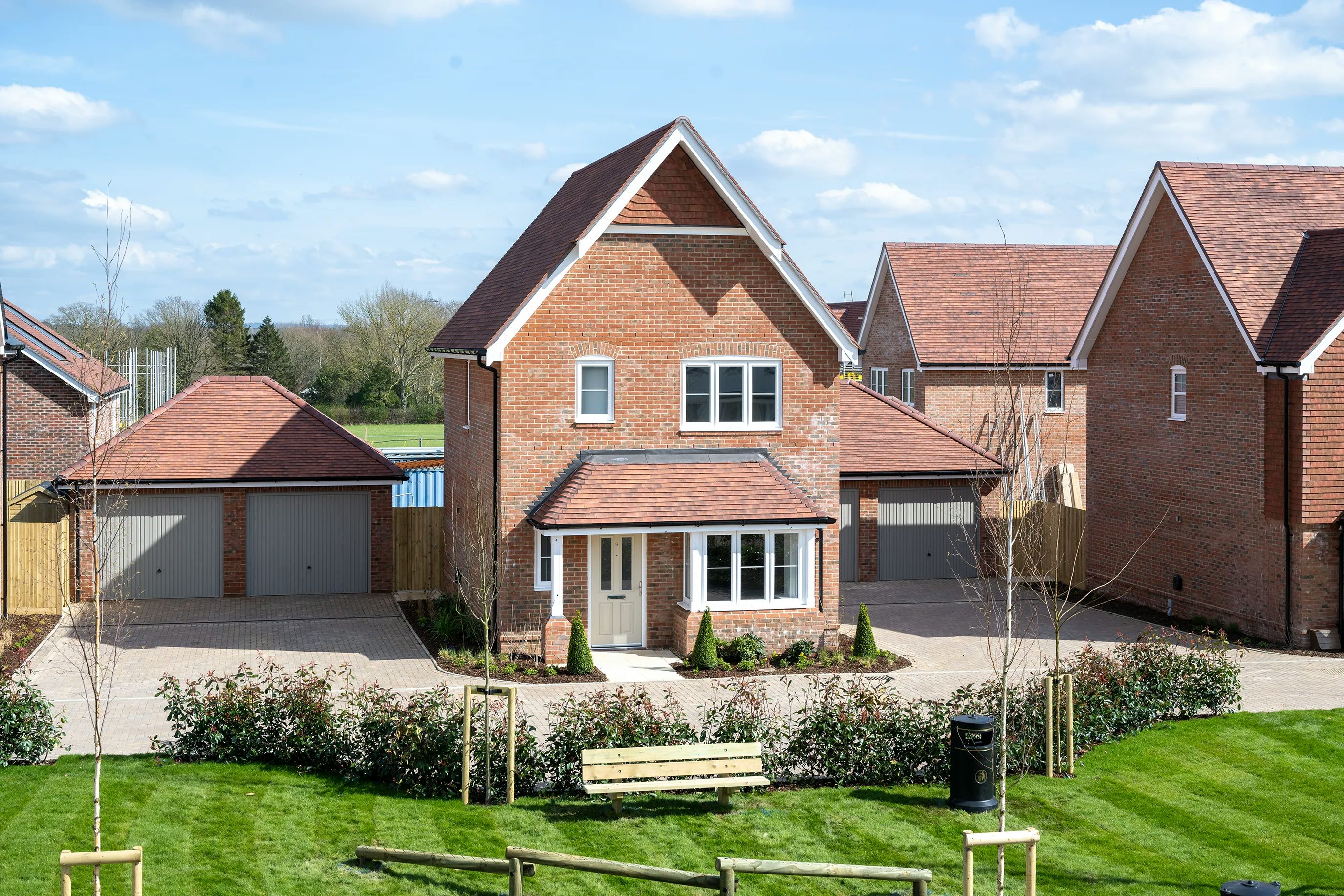 A modern, detached brick house with a pitched roof and double garage. The landscaped front yard features shrubs and a small tree. In the foreground, a grassy communal space includes benches and a trash bin. The sky is partly cloudy.