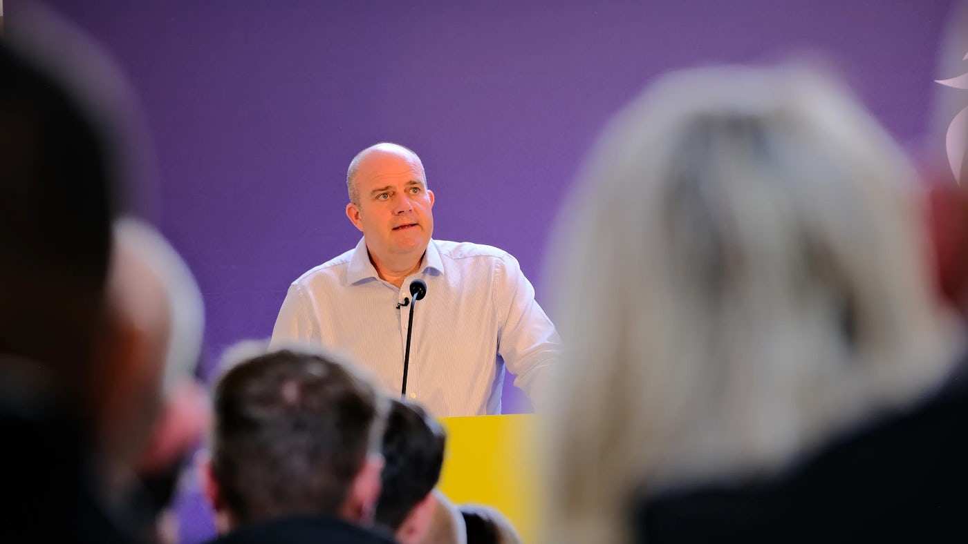 A person stands at a yellow podium, speaking into a microphone, with a purple backdrop behind them. They wear a light shirt adorned with a circular pin or badge. Several audience members are visible from behind in the foreground, facing the speaker.