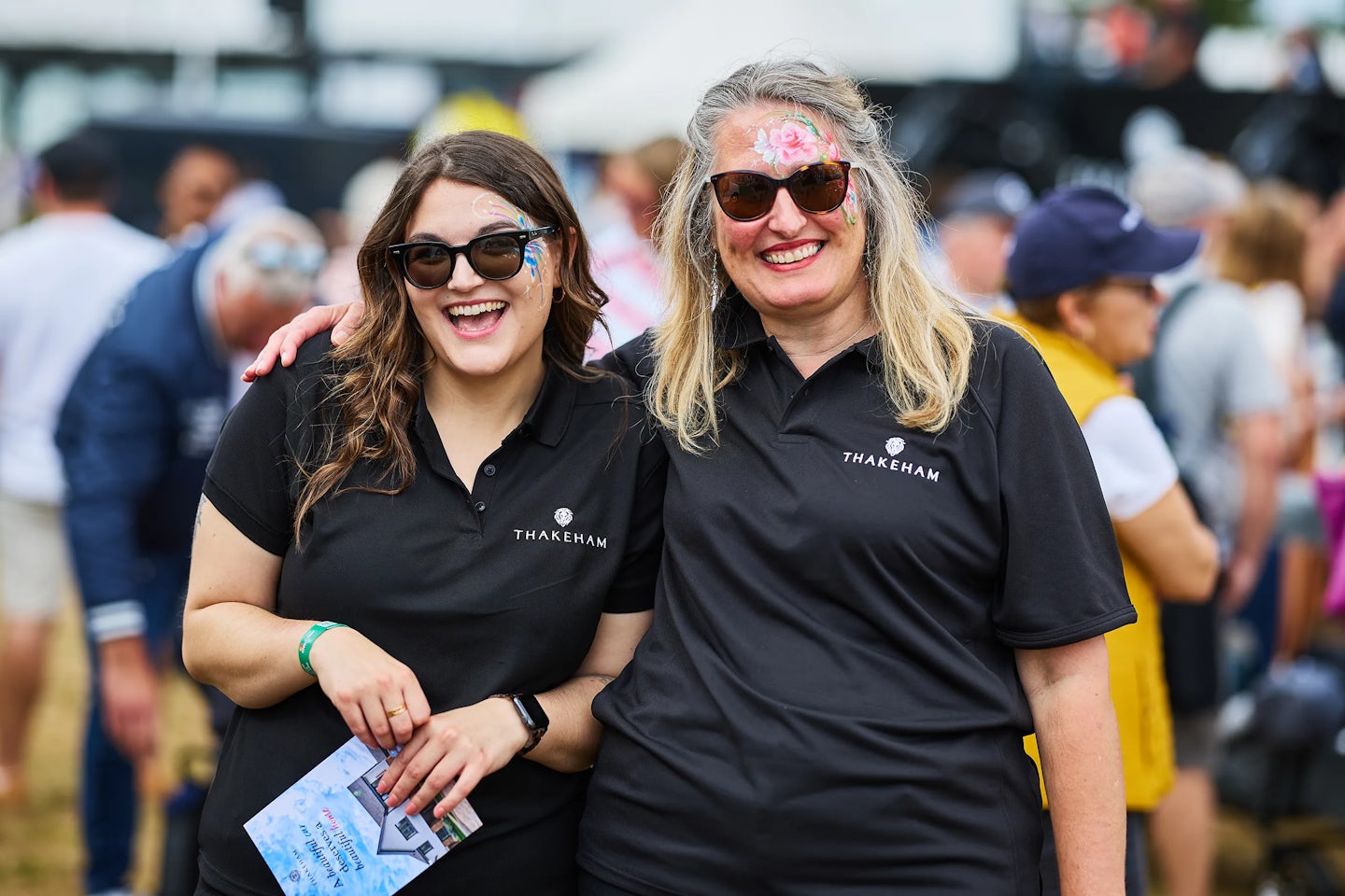Two individuals wearing black polo shirts embroidered with the “THAKEHAM” logo stand side by side at what appears to be a public event. One person holds an open brochure in both hands, suggesting they’re sharing information or engaging with attendees. A softly blurred crowd fills the background, reinforcing the sense of a busy, community-focused gathering. The overall atmosphere is professional and approachable, emphasising brand presence and outreach.