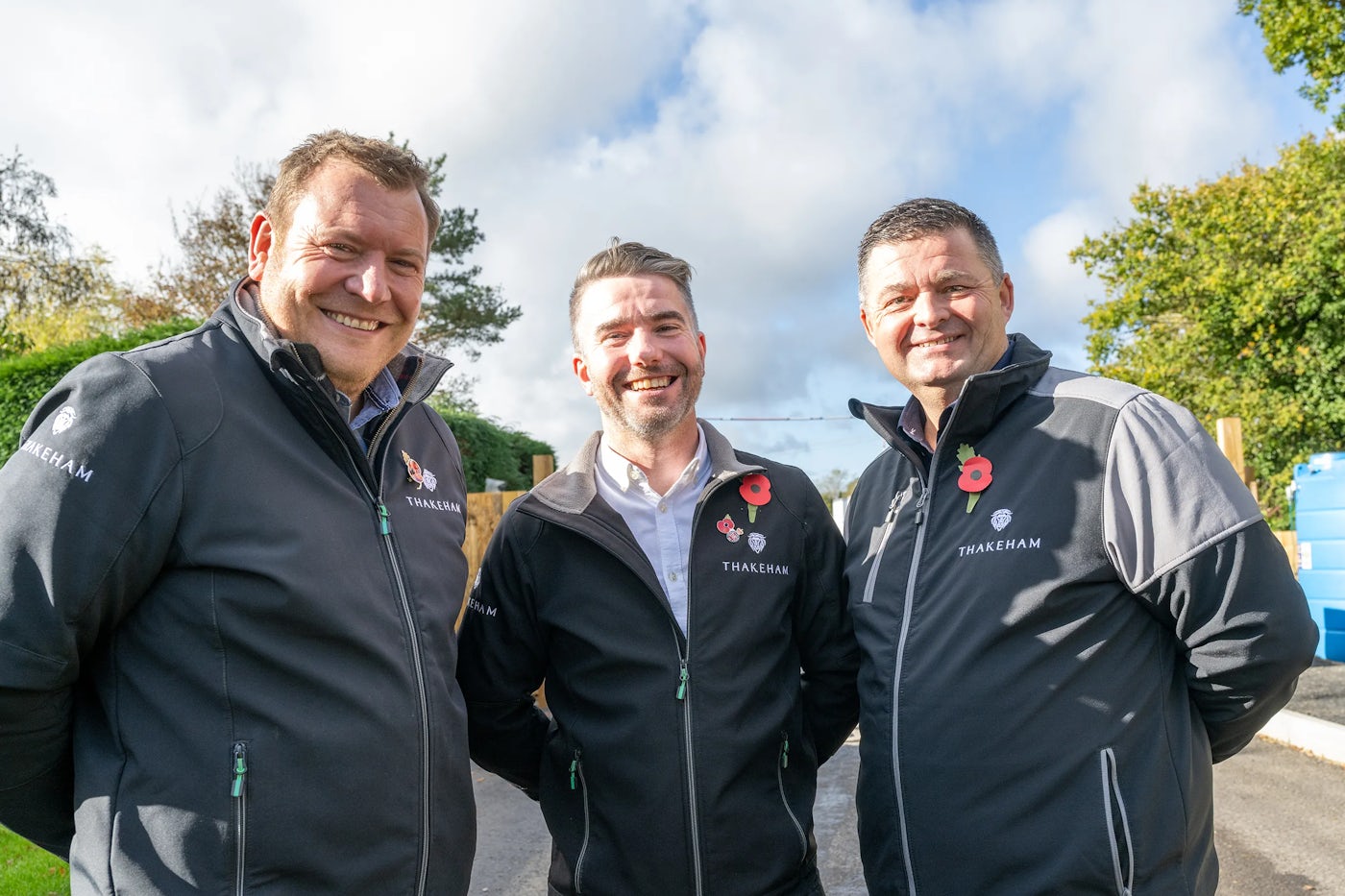 Three individuals stand close together on a paved path outdoors, framed by trees and shrubbery. Each wears a black THAKEHAM-branded jacket with the logo visible on the left chest and sleeve, and a red poppy pinned to their chest, suggesting a commemorative event—possibly for Remembrance Day. The overall tone is respectful and unified, evoking a sense of team spirit and shared purpose in a natural setting. Would you like to refine this for a particular platform or purpose—like internal communications, social media, or an accessibility context? I'm happy to tailor it.