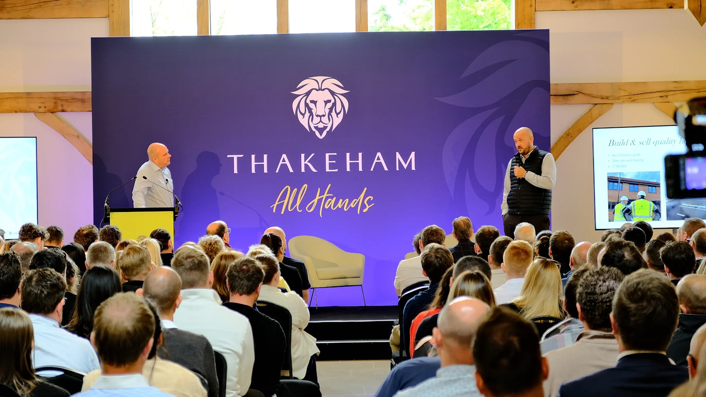 Indoor conference with a full seated audience facing a stage. Two speakers stand in front of a large screen and branded backdrop reading “THAKEHAM All Hands” with a lion emblem. One presenter stands at a podium on the left; the other speaks into a microphone on the right. A screen to the side displays the text “Build & sell quality” alongside a supporting image.