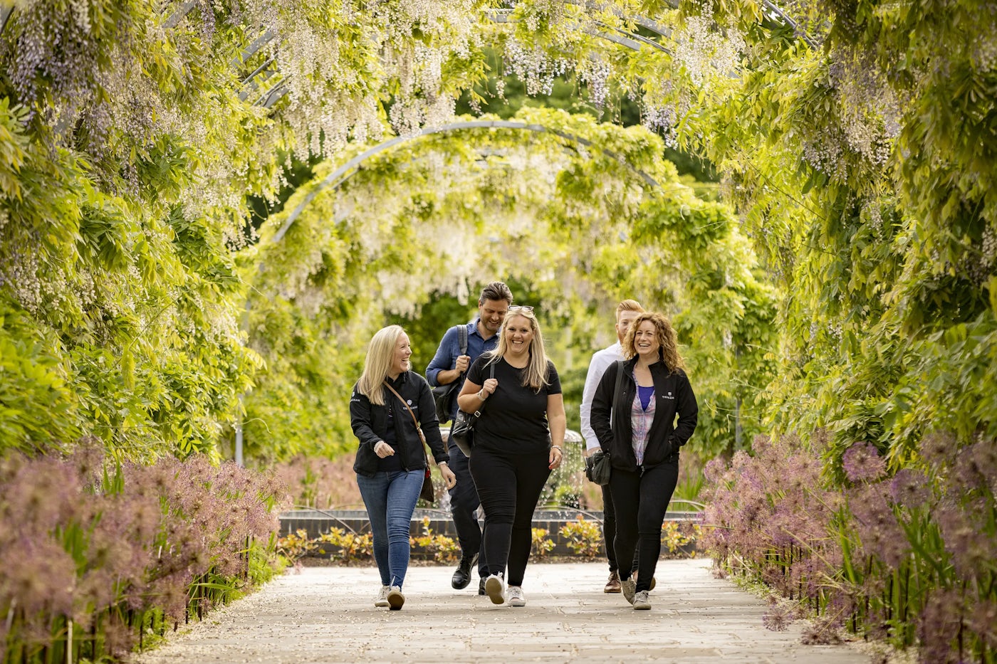 A group of five people walks along a garden path framed by arched trellises draped with flowers and greenery. The path is surrounded by vibrant, well-maintained plants, creating a peaceful, lush atmosphere ideal for a leisurely stroll. Soft natural light enhances the tranquil feel of the setting.