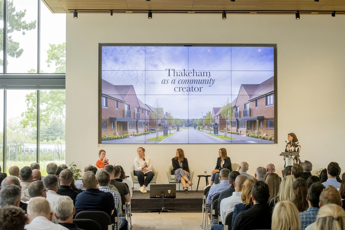 A modern conference room with four panelists seated on a stage in front of a large screen. The screen displays the text “Thakeham as a community creator” alongside an image of a suburban-style residential development. The audience, seated in rows, listens attentively. The space features tall windows that fill the room with natural light, creating an open, professional atmosphere.