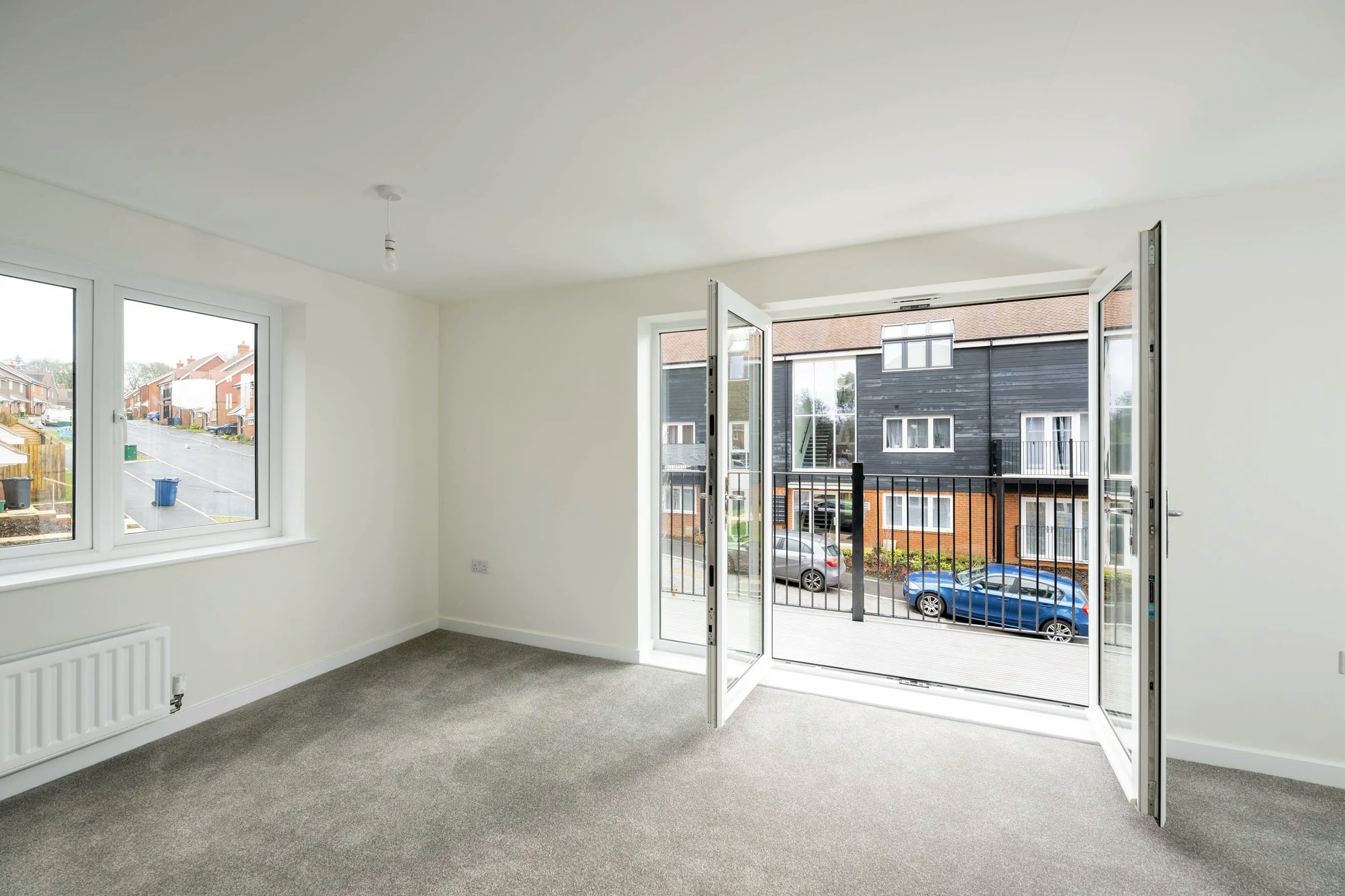 Bright, unfurnished room with pale walls and a soft grey carpet. Two windows on the left allow natural light to stream in, and double glass doors at the back lead to a small balcony overlooking a quiet residential street with parked cars and brick houses.