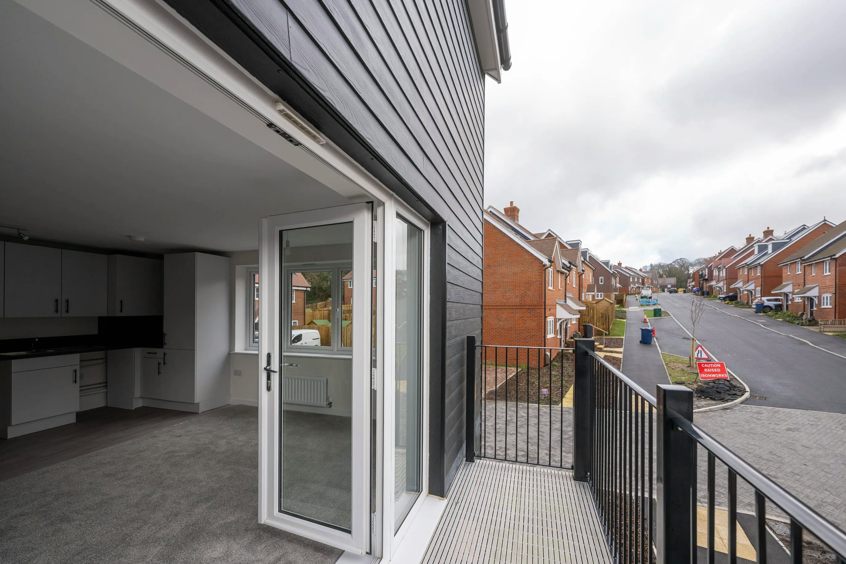 View from inside a modern house looking out through an open glass door to a balcony with a metal railing. The interior includes a compact kitchen with white cabinetry, a black countertop, and a carpeted floor. Beyond the balcony, a quiet residential street is visible, lined with similar homes under an overcast sky.