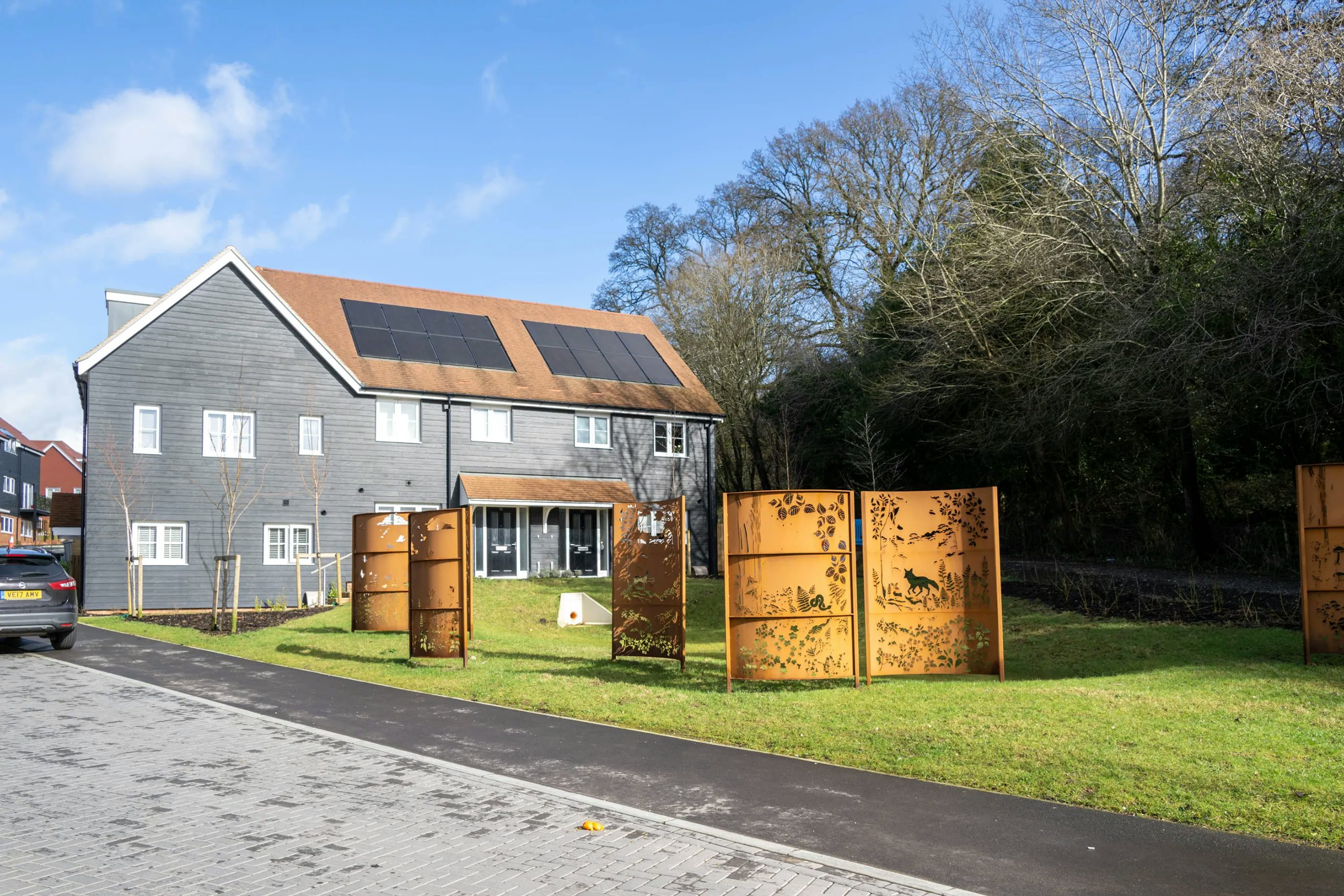 A modern two-storey residential building with grey cladding and white-framed windows and doors, topped with rooftop solar panels. In front, a row of decorative metal screens stands on a grassy verge beside a paved road. Each panel features intricate cut-out silhouettes of animals and plants, creating a whimsical, nature-inspired motif. The setting is bright and clear, with leafy trees and a blue sky in the background.