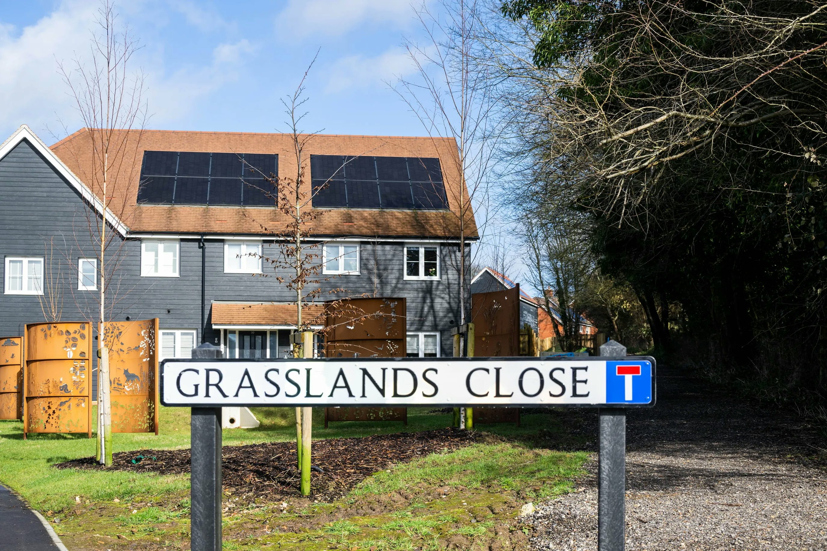 A street sign reading “GRASSLANDS CLOSE” stands in the foreground, accompanied by a no-through-road symbol. Behind it, a modern house with rooftop solar panels sits amid a grassy area, young trees, and a wooden fence. The background includes both leafy and bare trees beneath a clear sky with scattered clouds, reflecting a blend of seasonal transition and sustainable residential design.