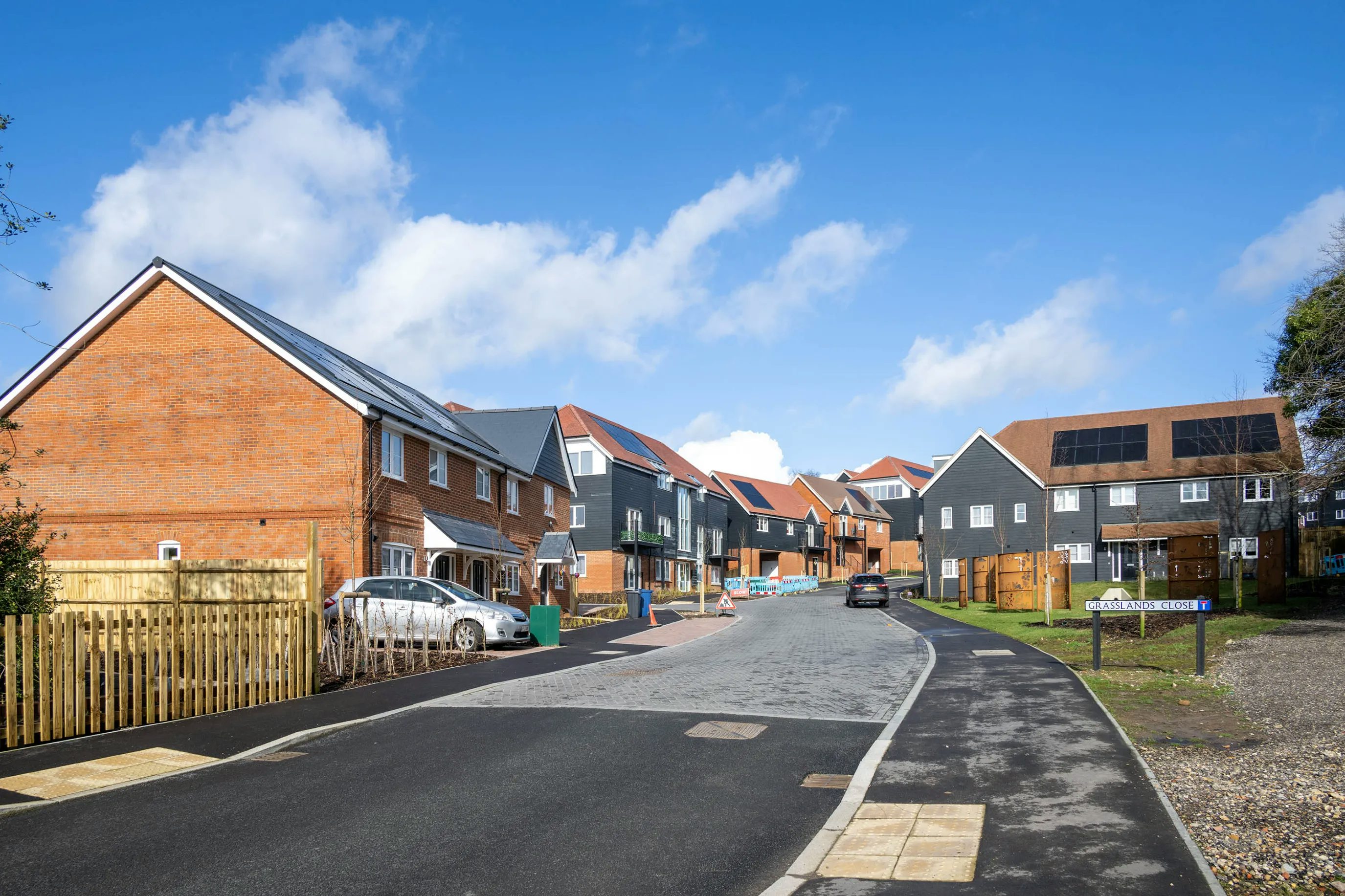 A residential street lined with newly constructed modern houses featuring a mix of red brick and dark grey siding exteriors with red-tiled roofs; cars are parked in driveways and along the curb; the street surface blends asphalt and cobblestone, with a paved footpath on one side; overhead, a bright blue sky with scattered clouds suggests a sunny day, highlighting a well-planned suburban development.