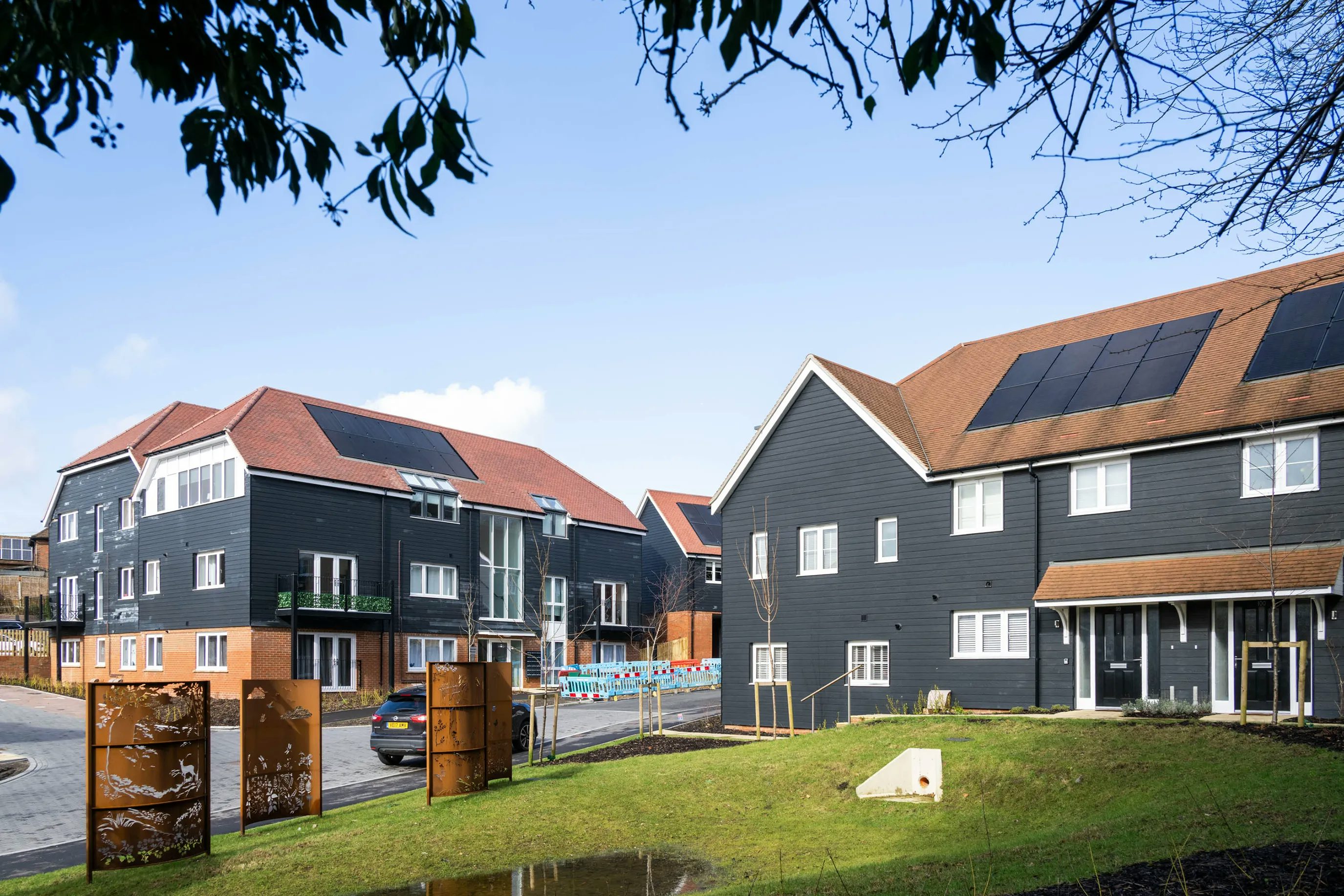 Modern residential buildings with dark façades, red roofs, and white-framed windows, one featuring rooftop solar panels. In the foreground, a parked car, grass verge, and small trees line the street, while construction barriers and decorative panels suggest ongoing development. A clear sky with light clouds completes the scene.