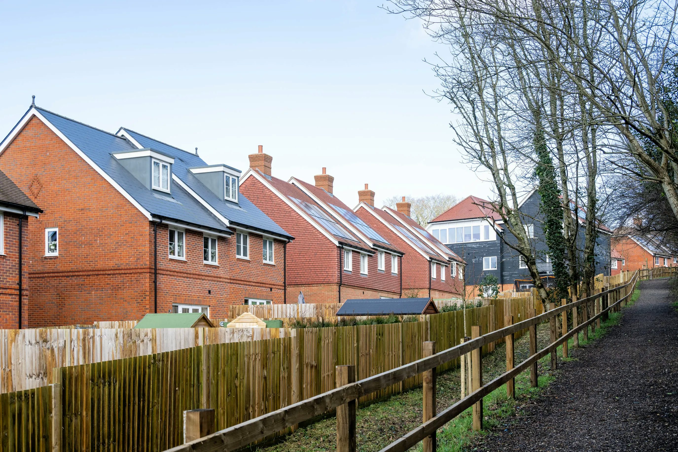 A row of modern brick houses with gabled roofs and dormer windows stands behind a wooden fence, bordering a light gravel path lined with leafy trees. The scene suggests a recently developed residential area, blending suburban tranquillity with contemporary design.