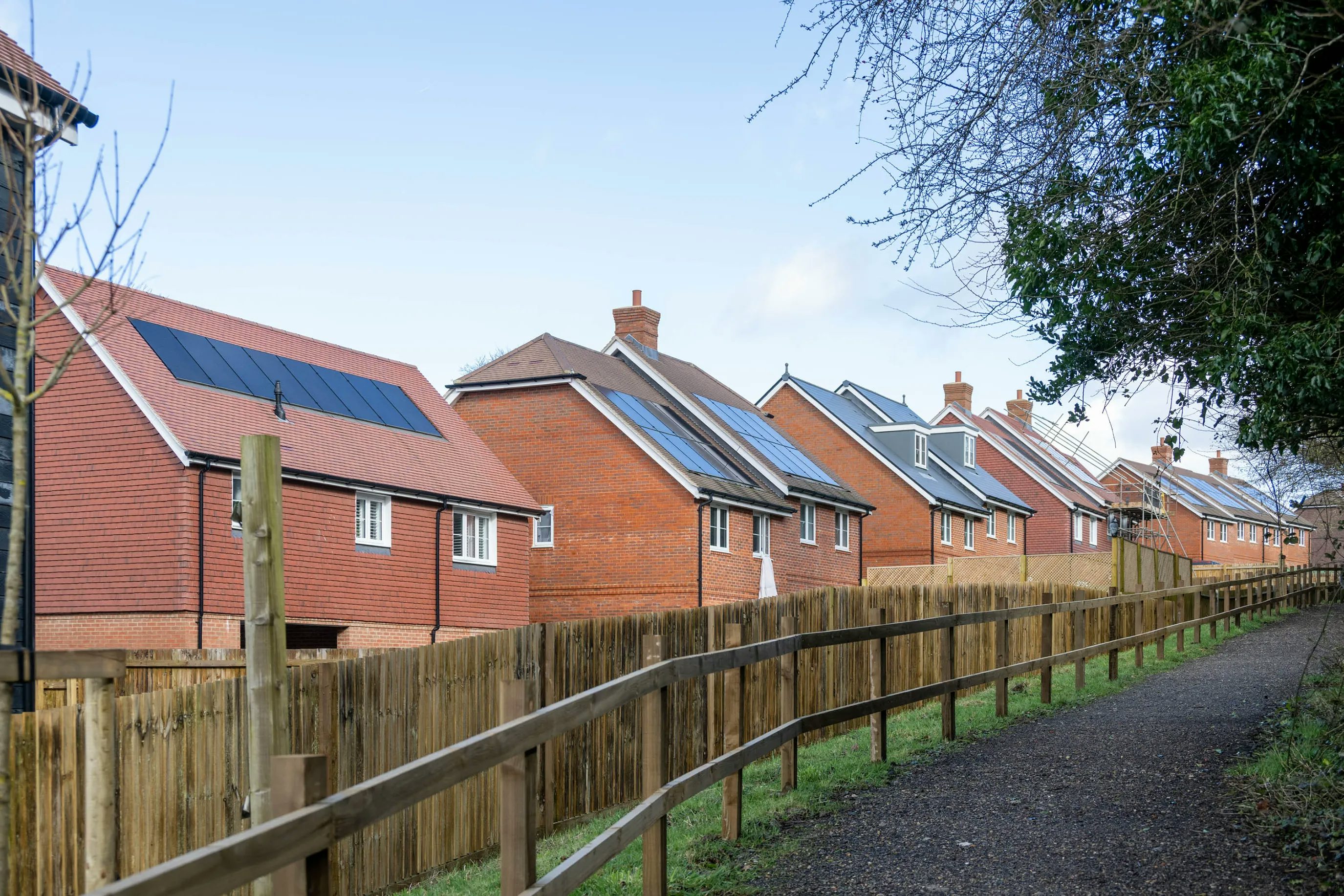 Row of modern red-brick houses with sloped roofs, each fitted with solar panels. A wooden fence lines a paved path bordered by grass and trees. The sky is mostly clear with scattered clouds, suggesting a calm residential setting focused on sustainability.