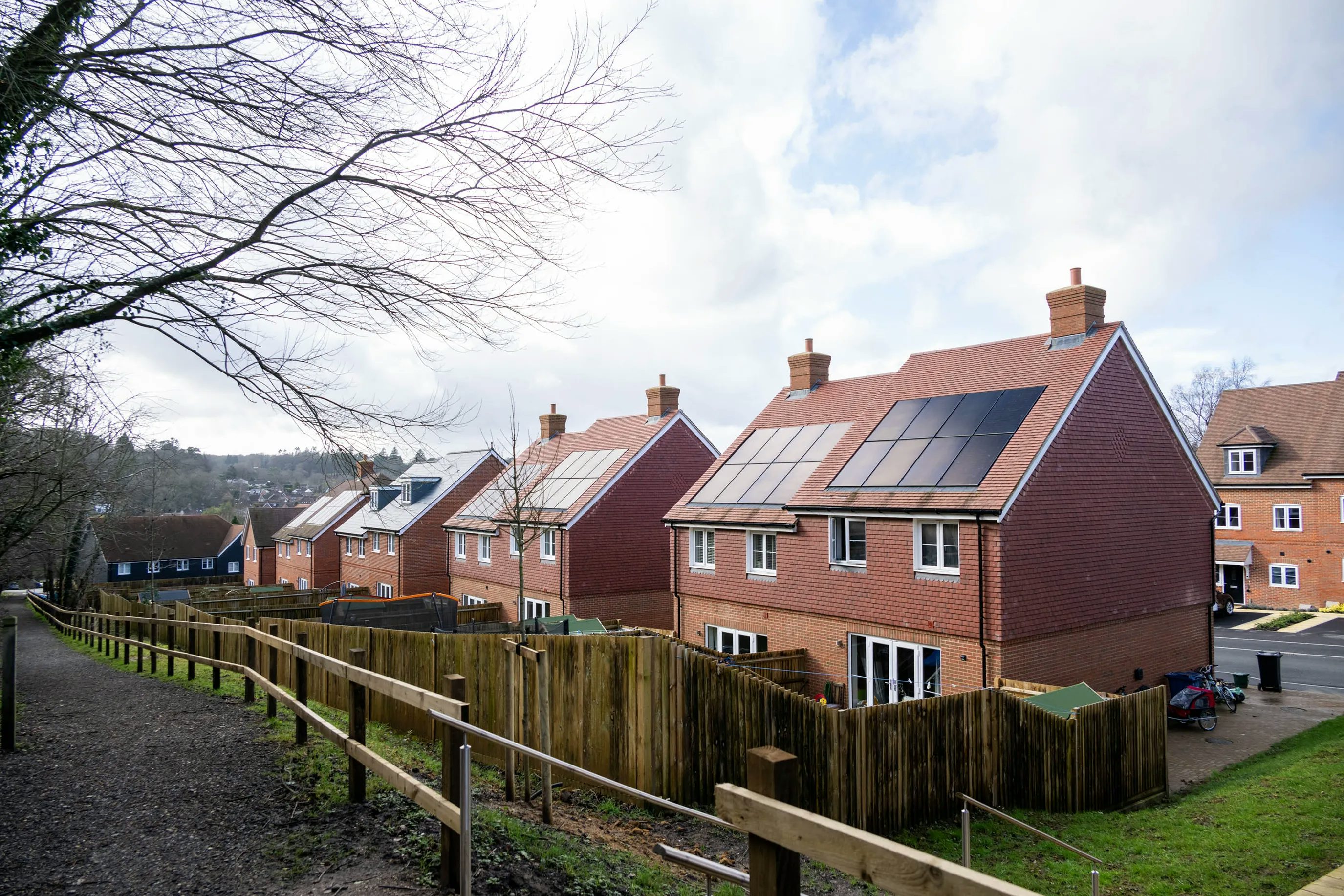 A row of modern two-story brick houses with solar panels on their pitched roofs, set along a fenced path with grass and trees on one side. The scene appears in a quiet suburban neighborhood under a partly cloudy sky, suggesting a focus on sustainability and residential design.