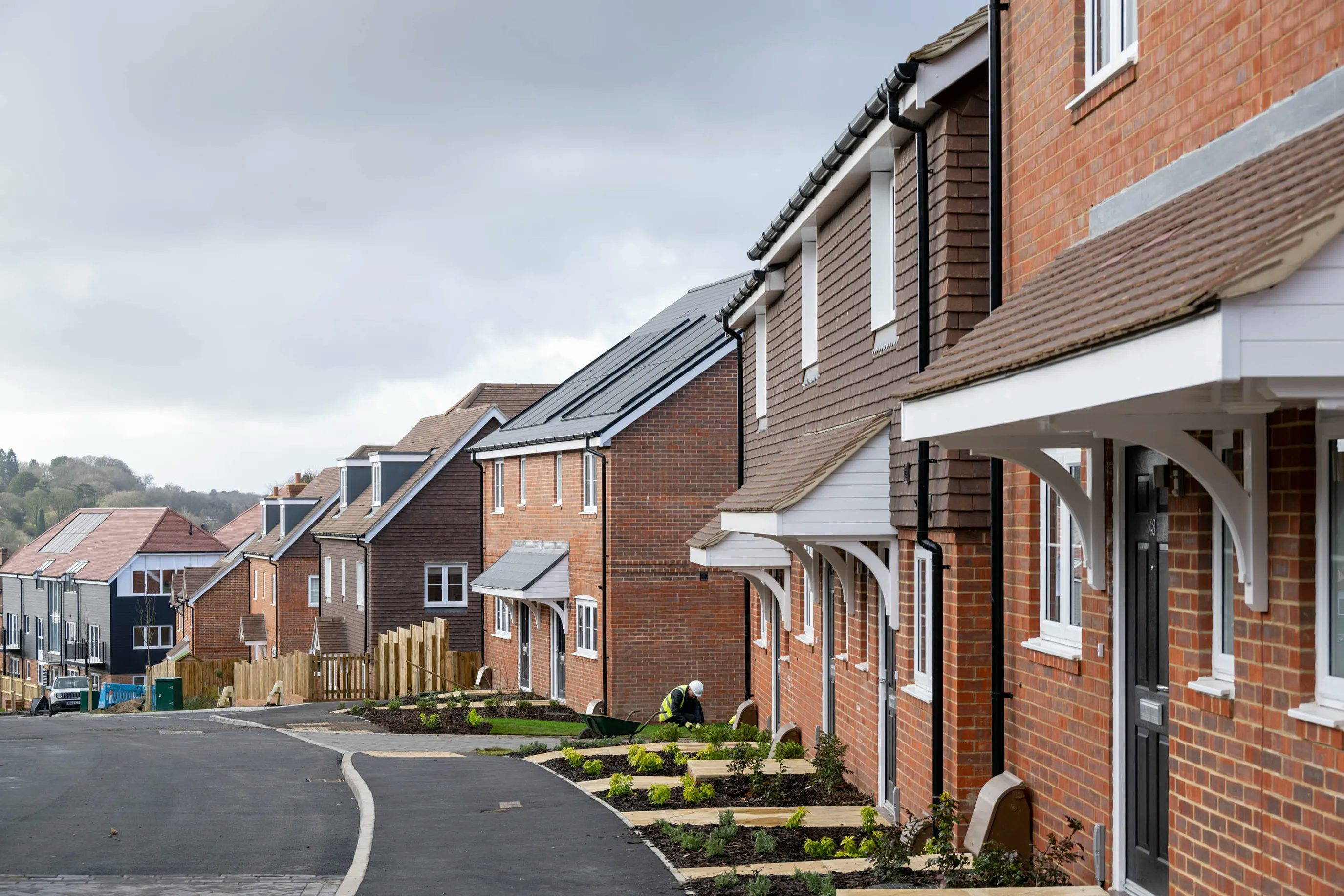 A row of newly built brick houses with white trim lines a gently curving paved street in a residential area. Each home has a small front garden with freshly planted shrubs. The sky is overcast, casting a soft grey light across the scene.