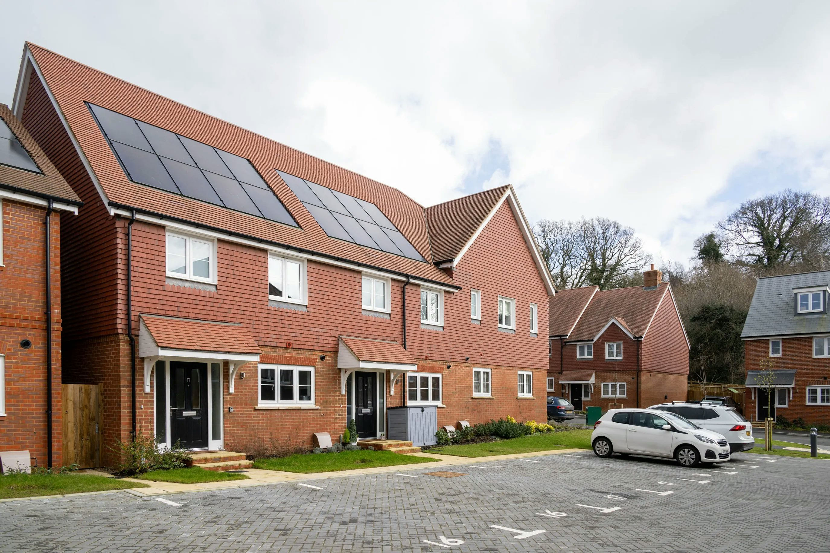 A row of modern, two-storey brick townhouses with pitched roofs, each fitted with solar panels. The homes feature small front gardens bordered by low hedges and are set along a paved parking area where several cars are parked. Mature trees and additional houses are visible in the background under an overcast sky, giving a calm, residential feel.