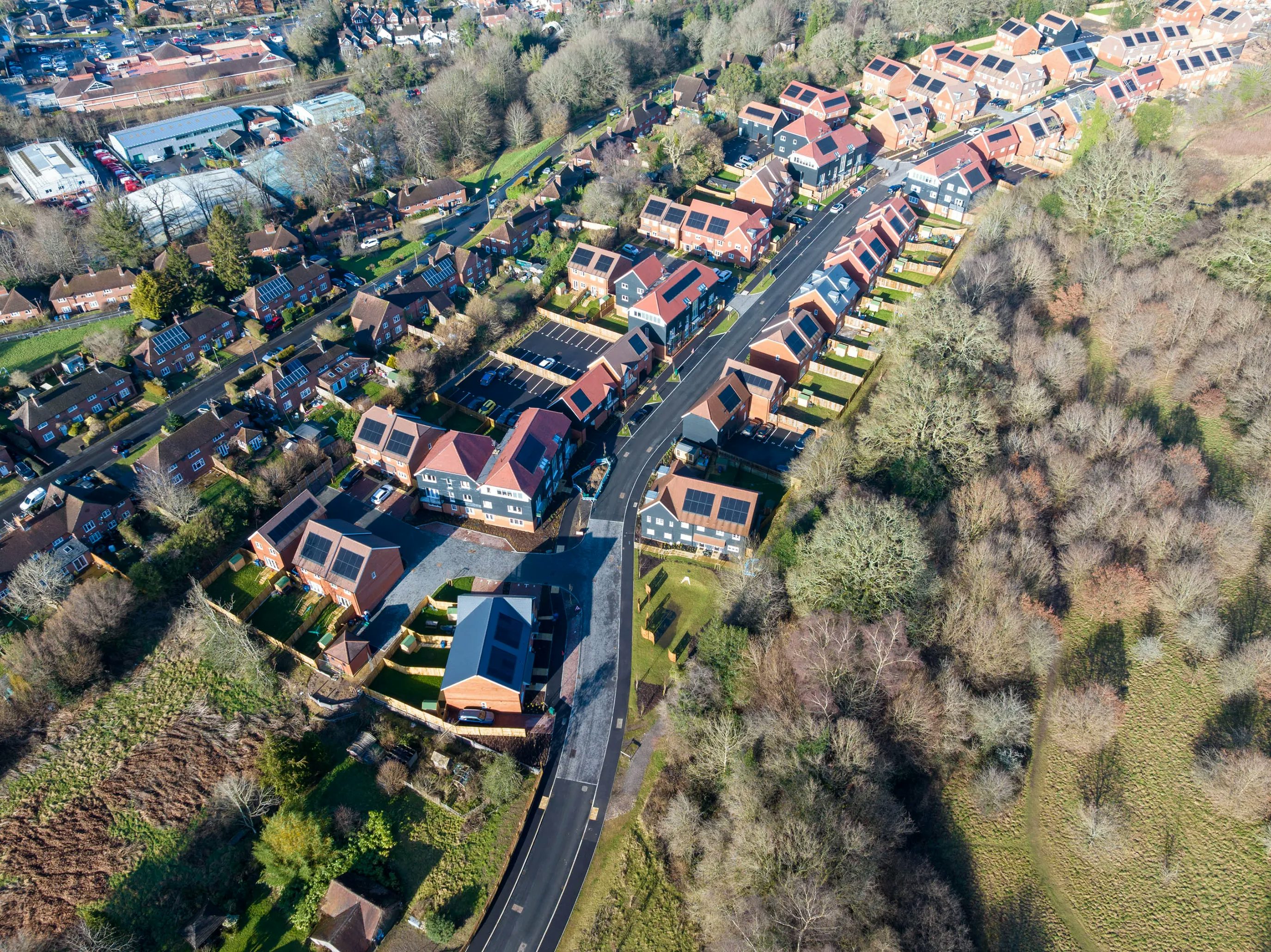 Aerial view of a neatly arranged residential neighbourhood featuring rows of houses with red roofs and solar panels, lining a curving main road that leads to a central roundabout. The scene is framed by abundant trees and greenery, highlighting the area's planned layout and suburban character. Natural light casts soft shadows across the rooftops and streets, creating a calm, orderly atmosphere.