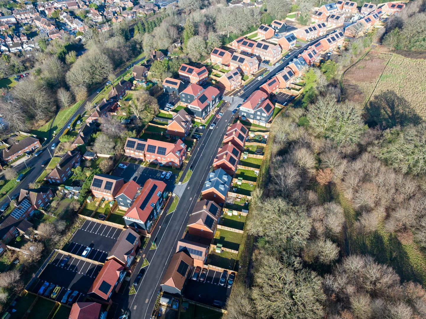 Aerial view of a neatly arranged residential neighbourhood featuring rows of houses with red roofs and solar panels, lining a curving main road that leads to a central roundabout. The scene is framed by abundant trees and greenery, highlighting the area's planned layout and suburban character. Natural light casts soft shadows across the rooftops and streets, creating a calm, orderly atmosphere.