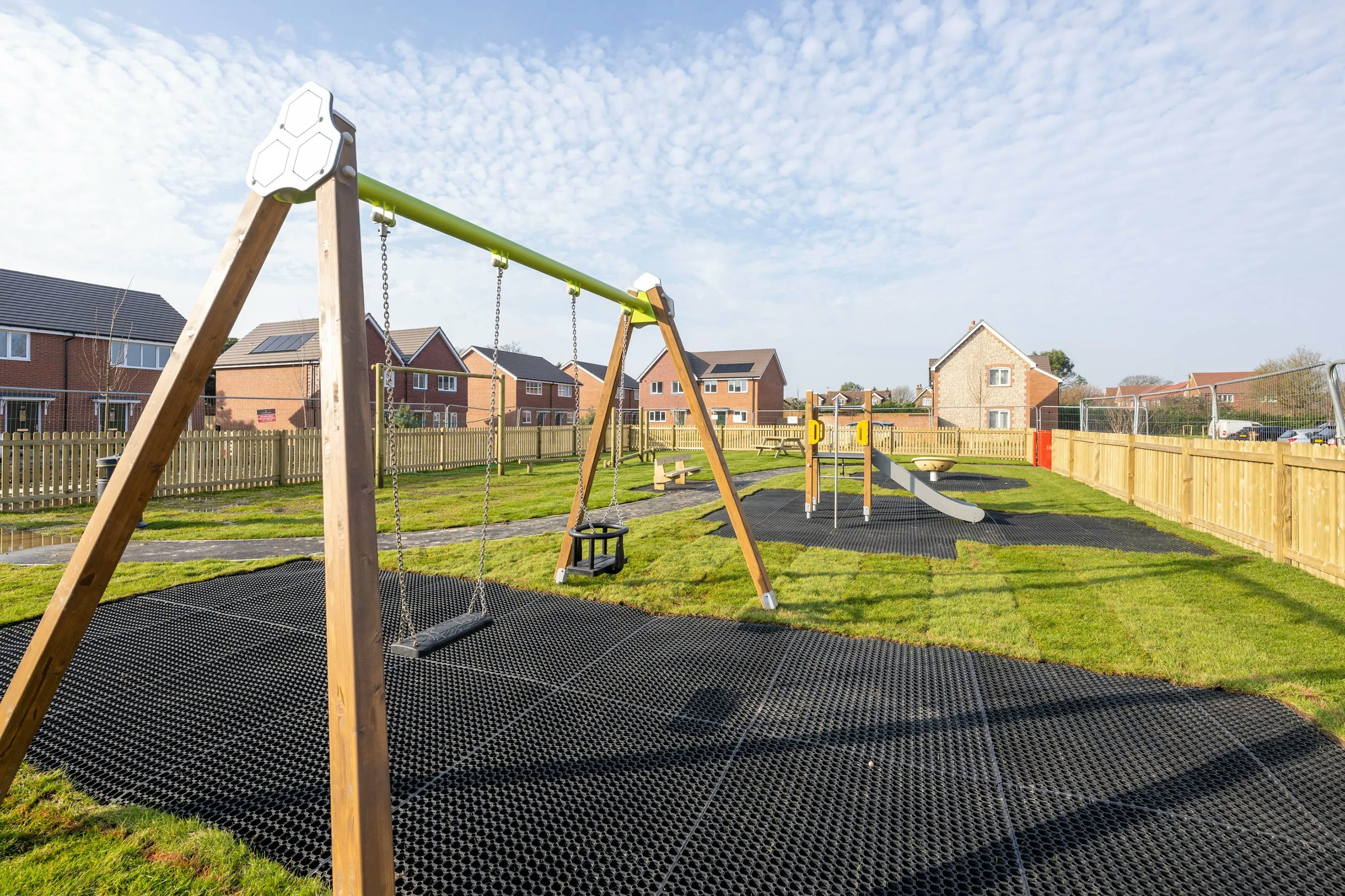 A well-maintained playground in a residential area, featuring a swing set with two swings, a slide, and other play equipment on safety mats. The space is enclosed by a wooden fence, with rows of brick houses and pitched roofs visible in the background under a partly cloudy sky.