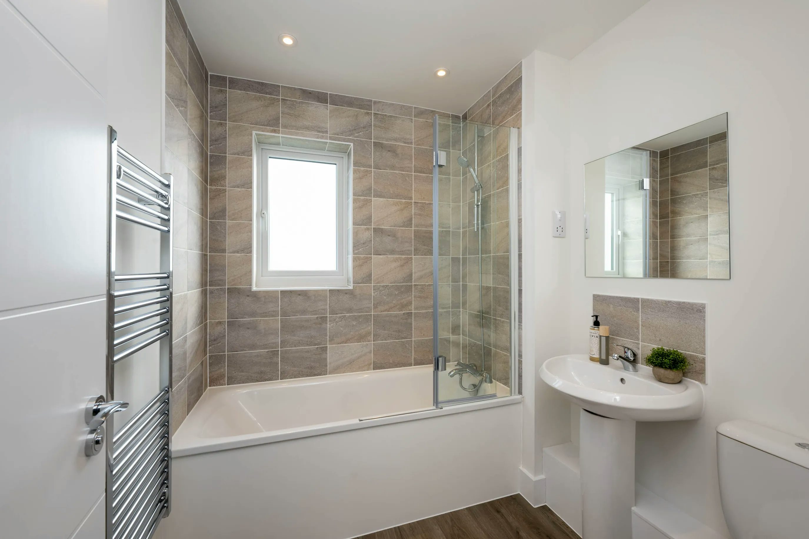 A modern bathroom with a clean, minimalist design featuring white walls and wood-effect flooring. A bathtub with a glass shower screen is set against a wall tiled in beige and brown, beneath a window that lets in natural light. A chrome towel radiator is mounted to the left of the tub. To the right, a white pedestal sink with a mirrored cabinet above it is positioned next to a toilet. The overall atmosphere is bright and contemporary.