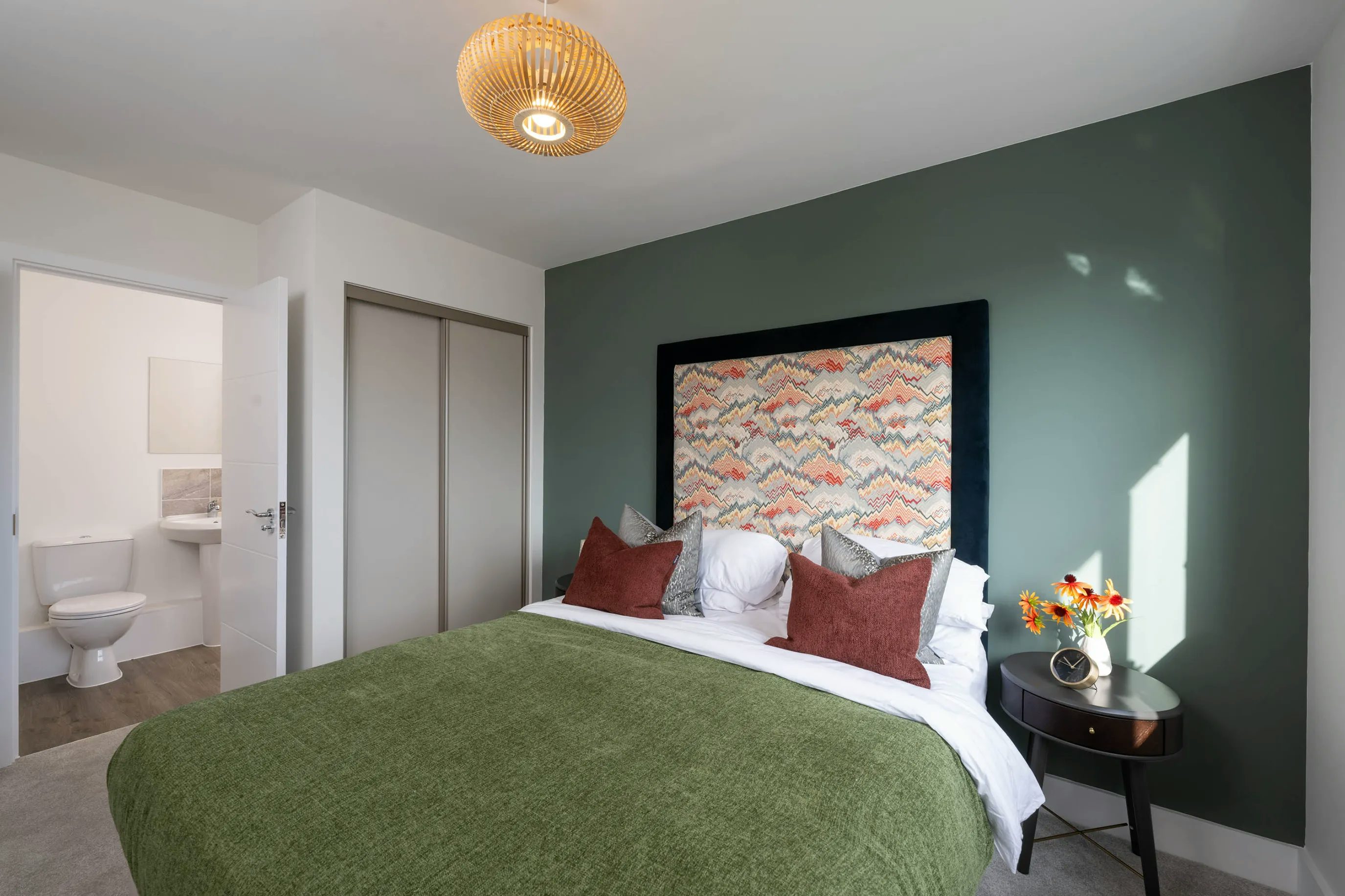 A modern bedroom with a green feature wall behind a double bed dressed in white sheets, a green throw, and layered pillows in brown and patterned covers. The headboard features a vibrant geometric design. A small round bedside table on the right holds a vase of flowers and a lamp. To the left, an open door reveals a bathroom with a visible toilet and sink. A closet with sliding doors lines one side, and a woven ceiling light fixture adds warmth and texture to the room.