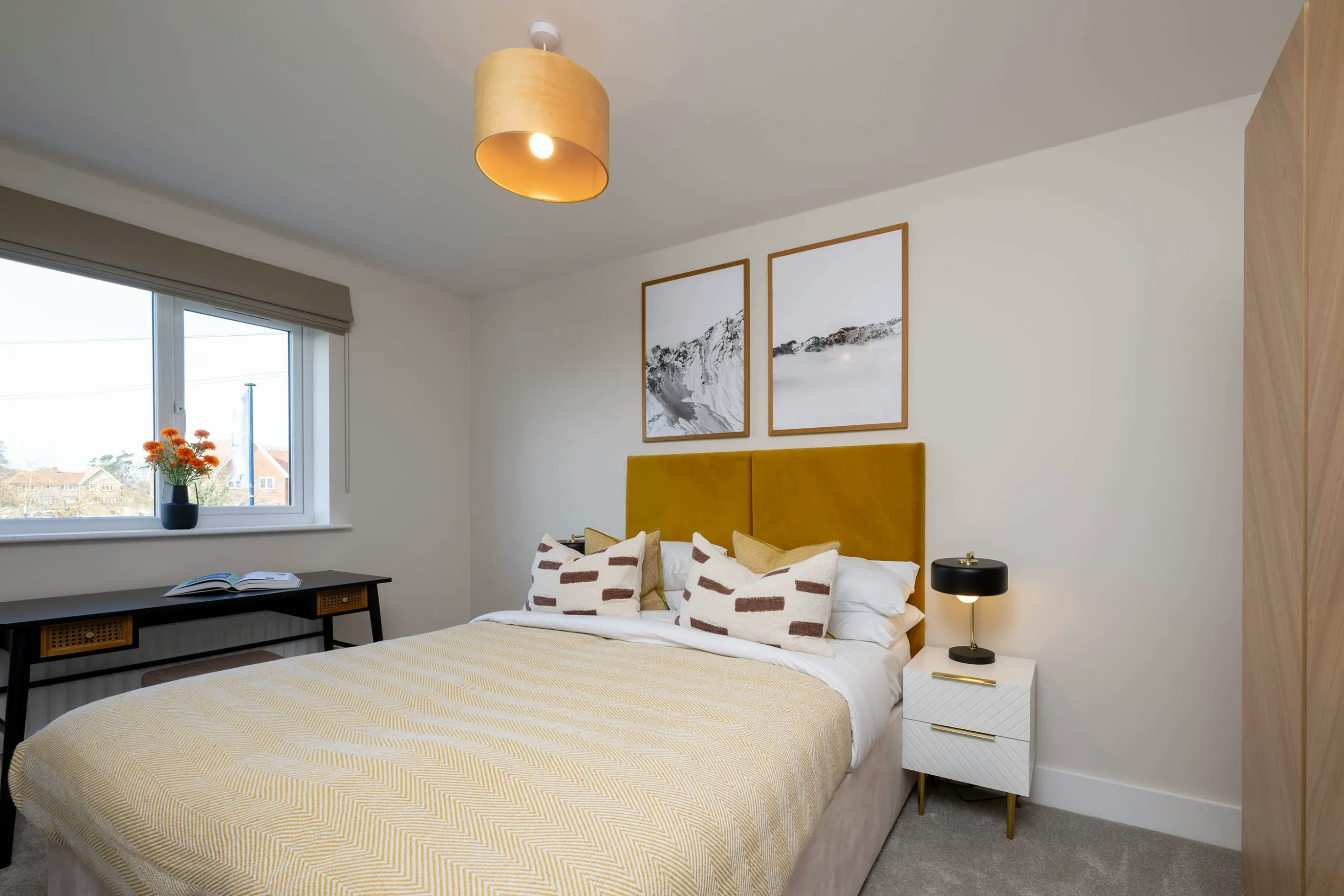 A modern, stylish bedroom with a mustard yellow and white colour palette. A double bed with a yellow headboard is neatly made with a matching bedspread and two geometric-patterned cushions. Above the bed hang two minimalist black-and-white framed prints. To the right, a sleek white nightstand with gold accents holds a black lamp. To the left, a tidy desk displays a vase of orange flowers, a book, and papers. A window with a beige blind offers a view of rooftops and leafy trees, while a ceiling light with a yellow shade softly illuminates the room.
