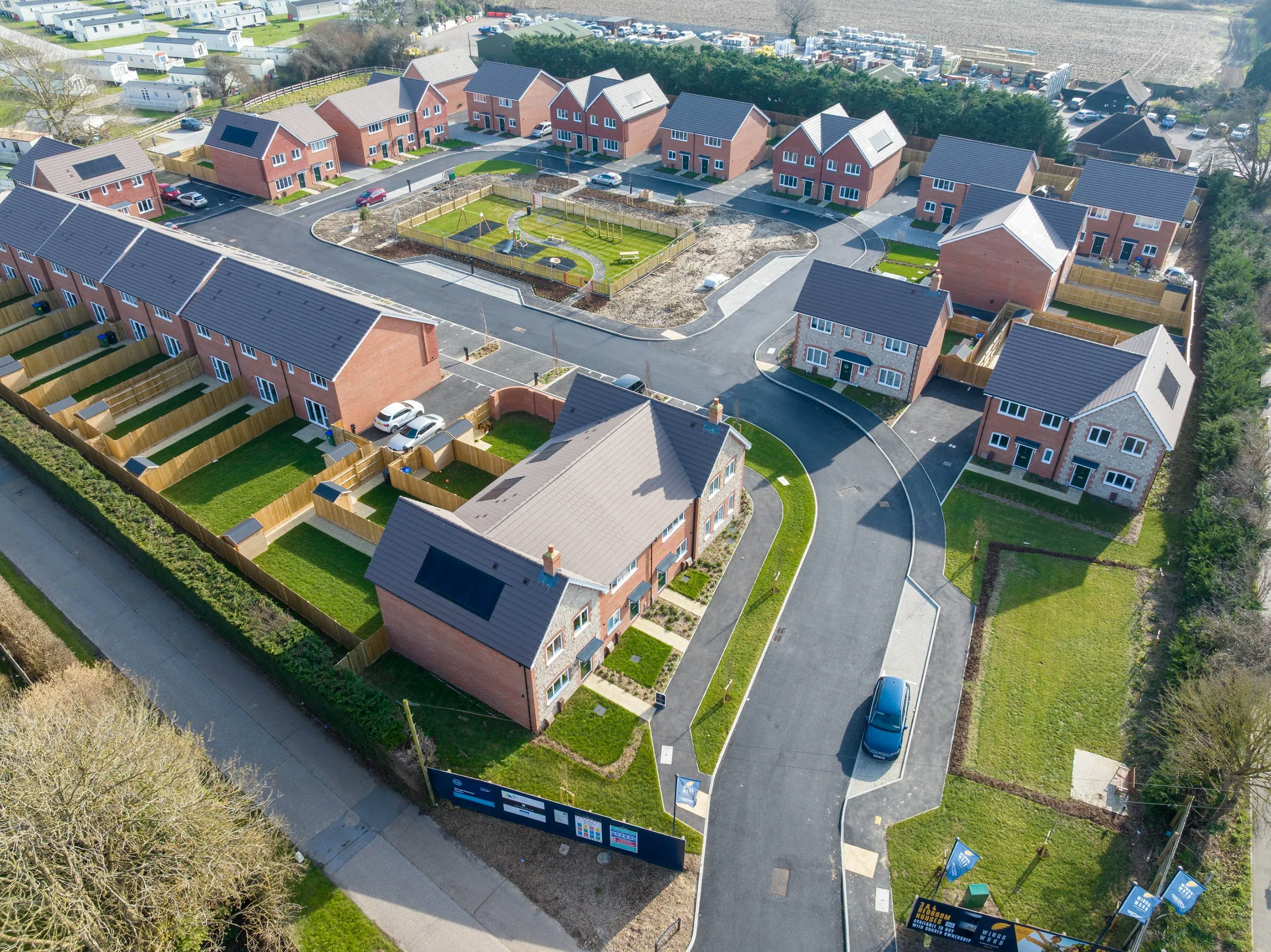 Aerial view of a newly built residential neighborhood featuring rows of red-brick houses with dark roofs, some fitted with solar panels. Homes are arranged around a central green space with a construction site indicating ongoing development. Paved roads, fenced gardens, and symmetrical layouts reflect modern urban planning. Would you like this refined to focus more on sustainability, community, or another specific theme? I’m happy to tailor it.