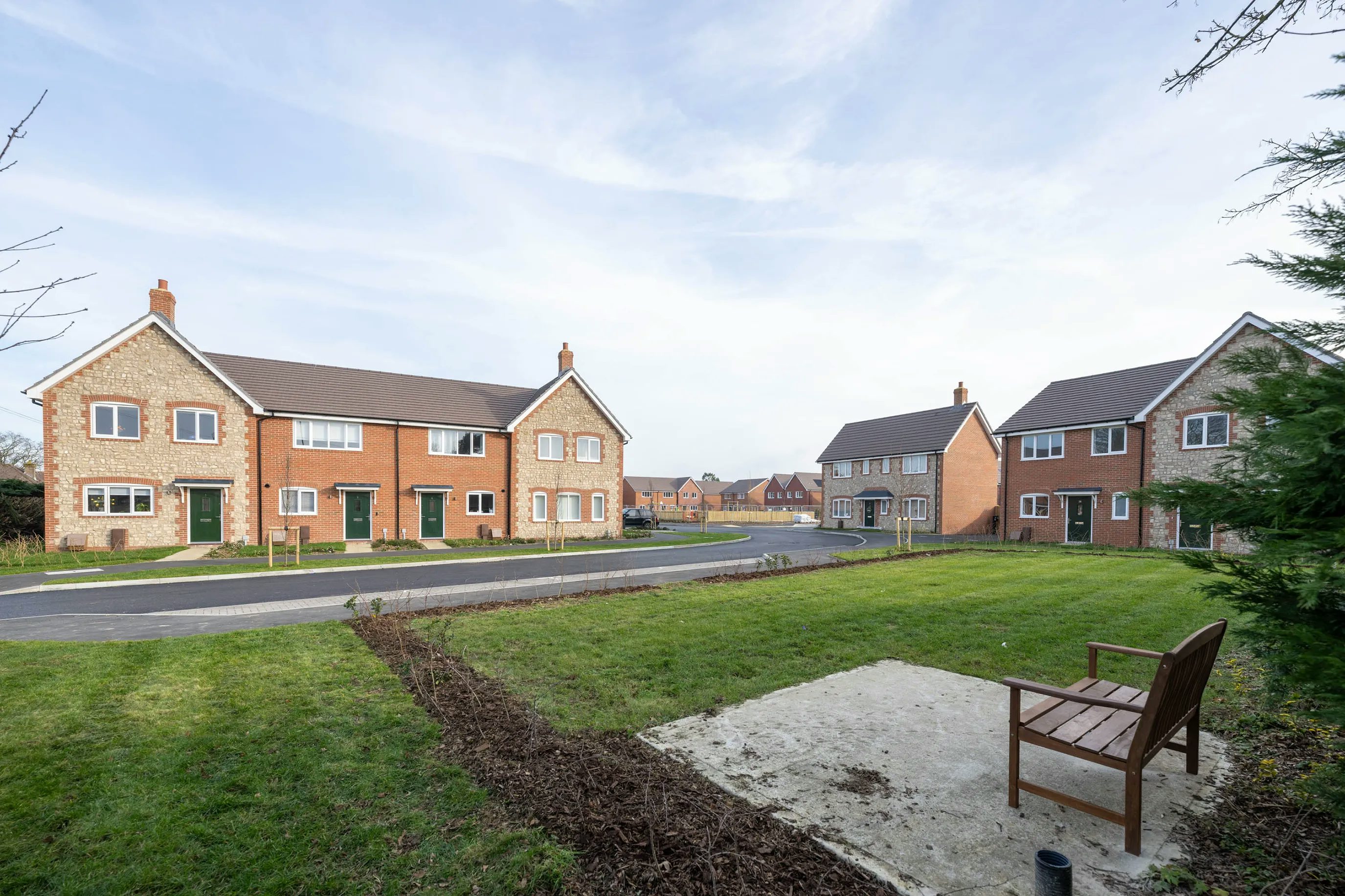 A newly developed suburban neighbourhood with two rows of modern brick houses facing each other across a quiet residential street. Each house features a pitched roof and small front garden. In the foreground, a grassy verge with a wooden bench set on a square concrete base adds a welcoming touch. The sky above is mostly clear with a few scattered clouds, enhancing the calm, well-kept atmosphere of the area.