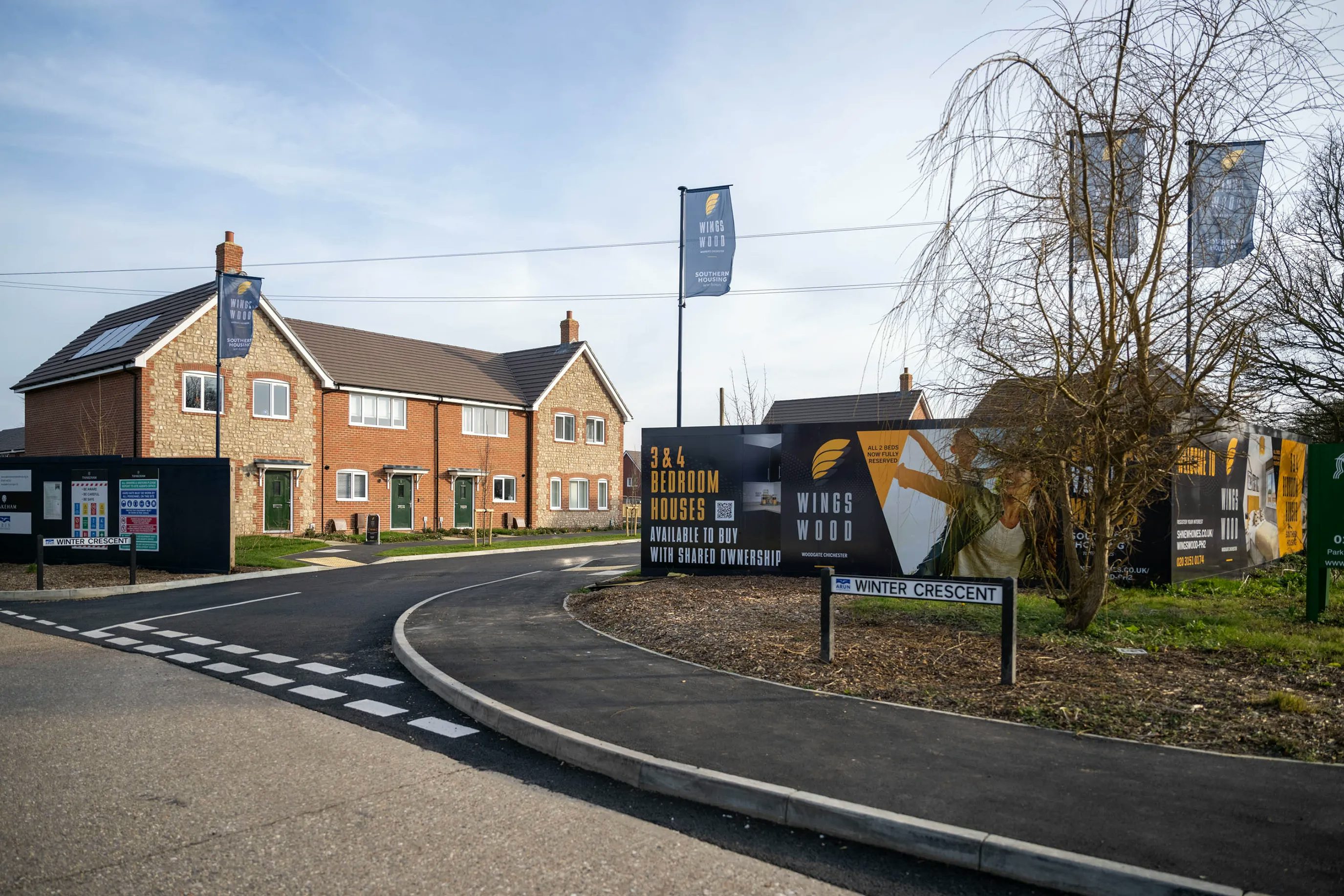 Newly built red-brick homes on a residential street named Winter Crescent, featuring green front doors and solar panels on pitched roofs. Prominent signs advertise shared ownership options for three-bedroom houses in the Wings Wood development. The paved street includes clean sidewalks and modern lamp posts under a bright sky.
