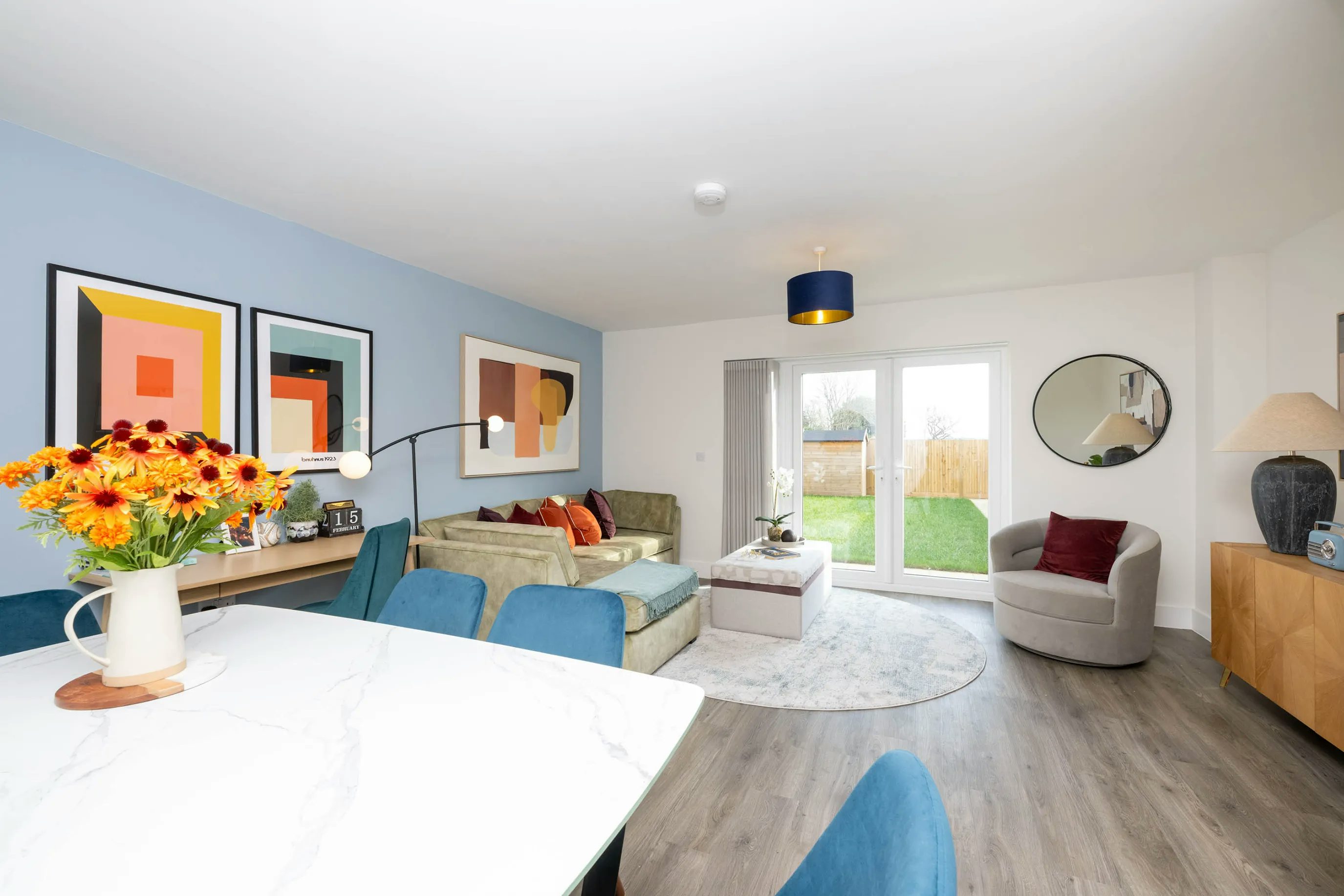 Modern living room with a blue feature wall displaying three abstract artworks above a beige L-shaped sofa accented with red and orange cushions. A round mirror hangs on the adjacent white wall. A grey armchair with a red pillow sits beside a round rug and rectangular coffee table. Full-height glass doors reveal a green garden beyond. On the right, a wooden sideboard holds a lamp and decor, while in the foreground, a white marble dining table with blue chairs features a vibrant bouquet of yellow and orange flowers. The space is bright with natural light and a blue-shaded ceiling lamp. Would you like to refine this for a particular platform or tone—more casual, brand-aligned, or accessibility-focused? Happy to iterate with you.