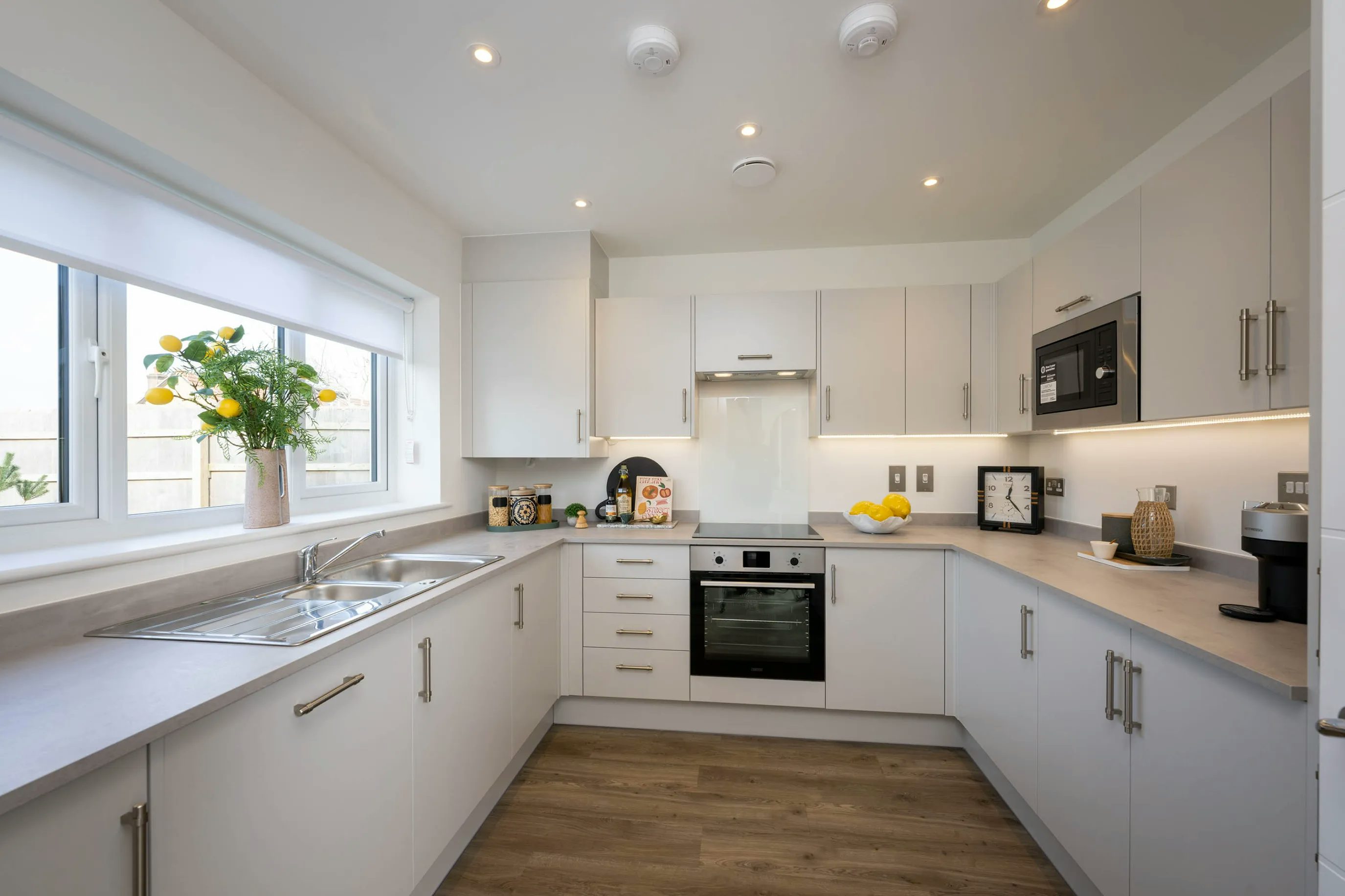 Modern kitchen with white cabinetry, sleek countertops, and stainless steel appliances, including a double sink beneath a window with a white blind. A coffee maker, decorative containers, and a bowl of lemons add colour to the clean, well-lit space with wooden flooring and recessed lighting.