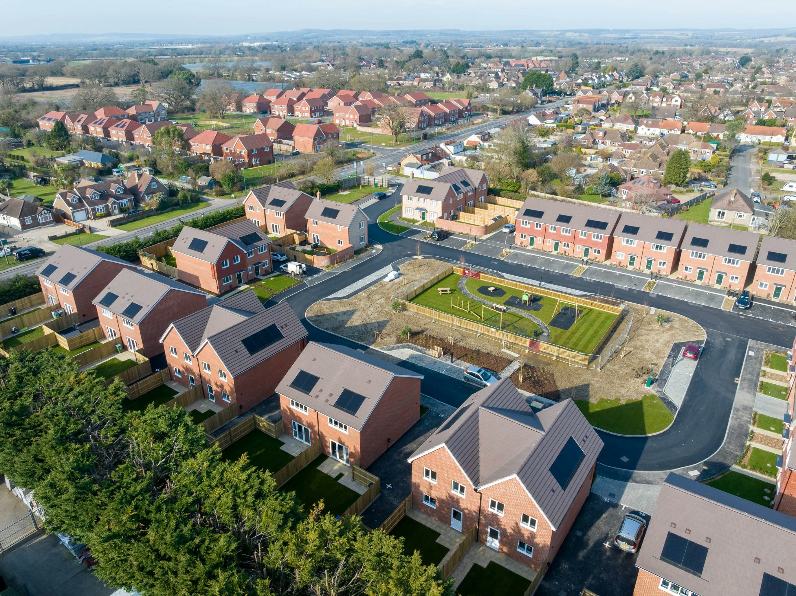 Aerial view of a modern residential neighborhood arranged in a planned layout. Brick-built houses with tiled roofs line curved roads and cul-de-sacs, some featuring solar panels. A landscaped central green space includes a circular path, garden beds, and a small playground. Surrounding trees and distant housing blend suburban living with accessible green areas, reflecting thoughtful urban planning and community design.