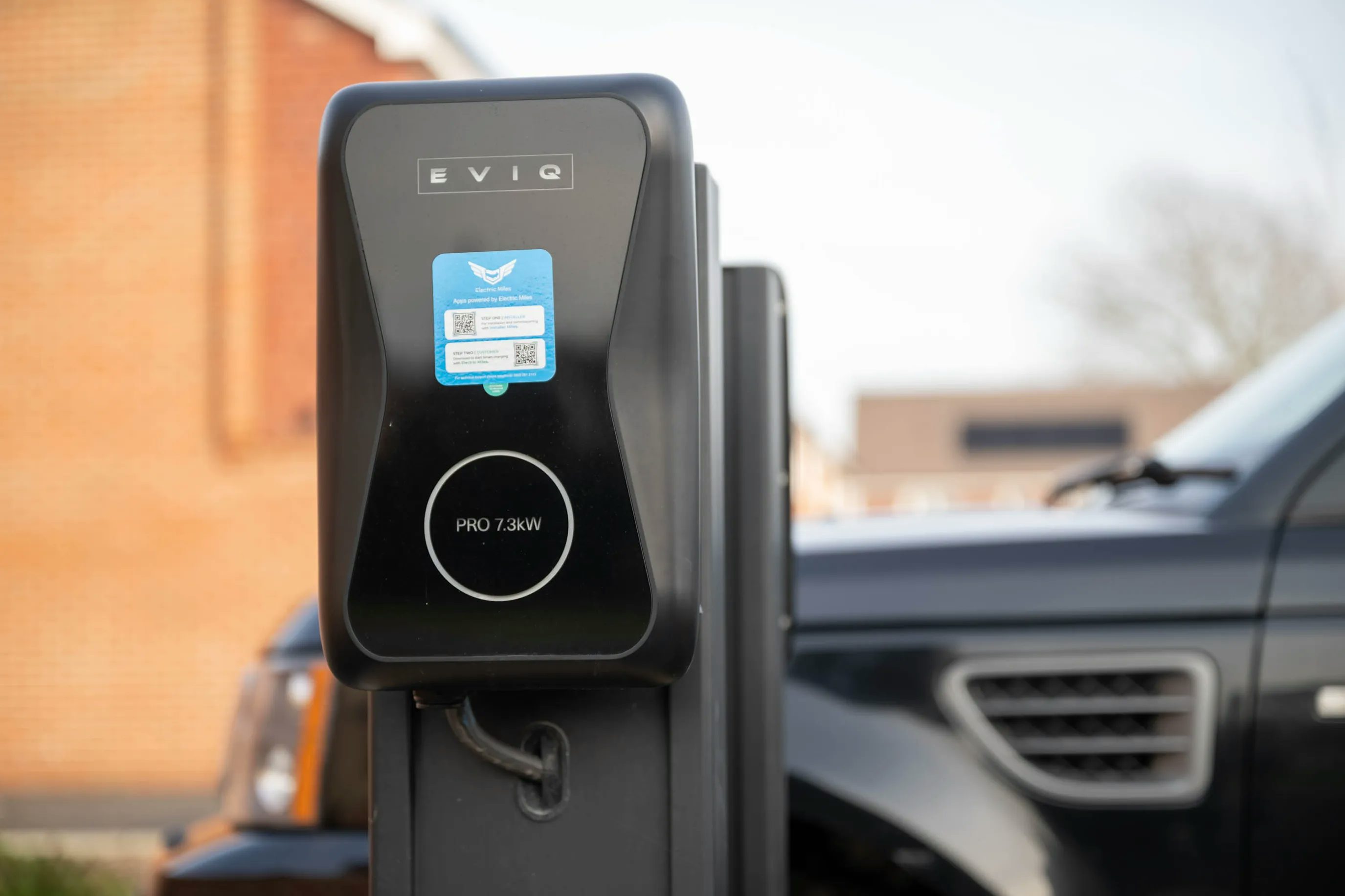 Electric vehicle charging station marked “EVIQ” with a “PRO 7.3kW” label, featuring a blue QR code sticker. A black car is parked nearby, and a brick building forms the background, reflecting a modern approach to sustainable transport infrastructure.