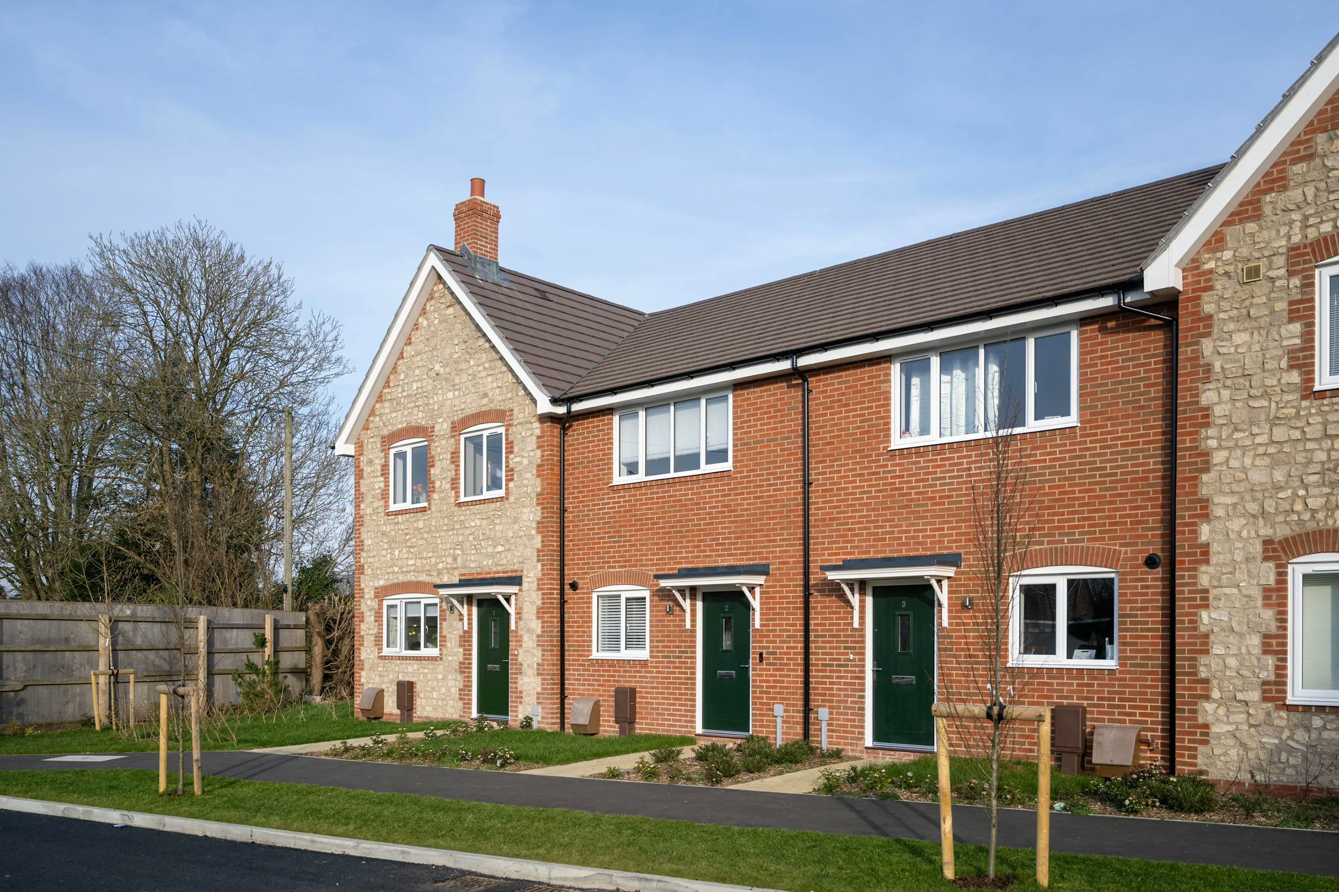 A row of contemporary terraced houses with a mix of red brick and pale stone facades. Each two-storey home features a green front door, white-framed windows on both levels, and a small landscaped front garden. A shared paved pathway runs in front of the houses, bordered by neatly cut grass and young trees. The scene is set on a bright, sunny day in a suburban neighbourhood.