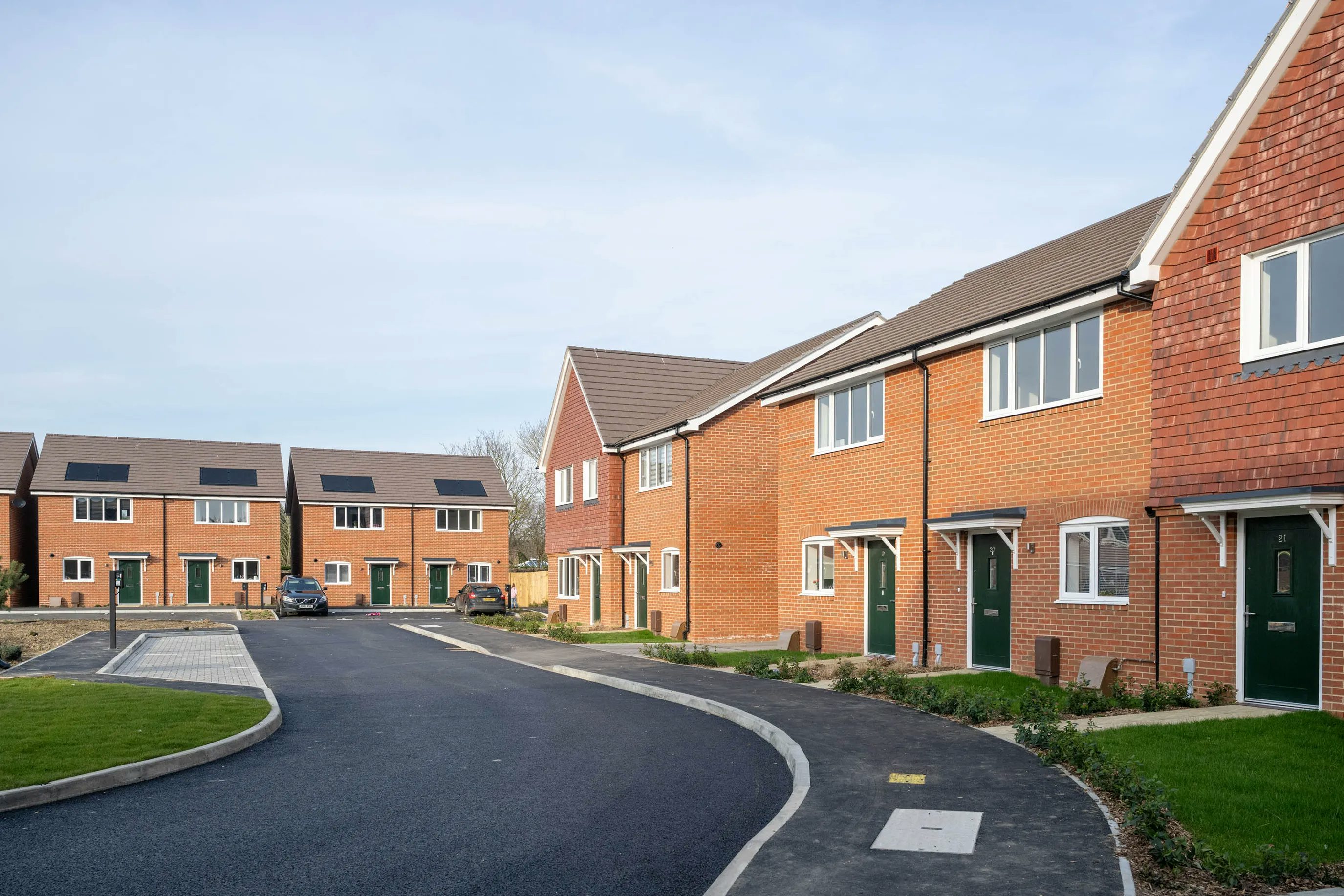 A sunny residential street featuring newly built brick houses with green doors and white-framed windows. The paved road curves gently left, bordered by a sidewalk and patches of grass. A few cars are parked outside the homes, under a clear blue sky, evoking a calm, suburban atmosphere.
